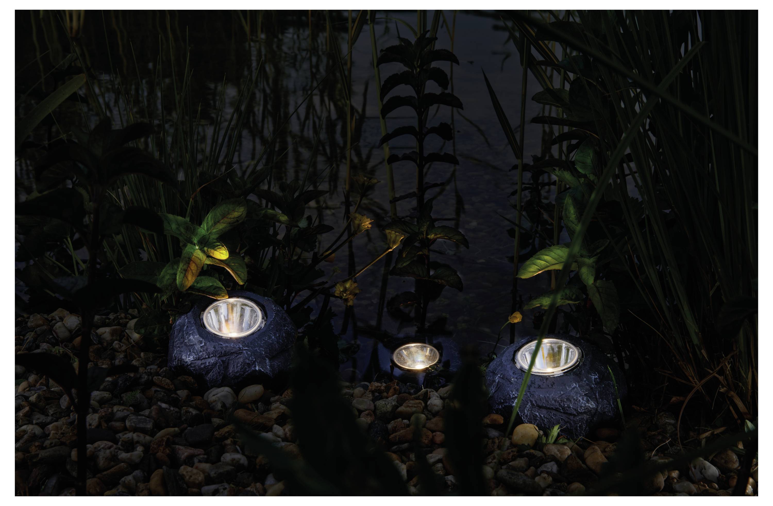Trois lampes solaires ressemblant à des rochers illuminent un chemin de jardin bordé de plantes et de galets la nuit, créant une atmosphère sereine.