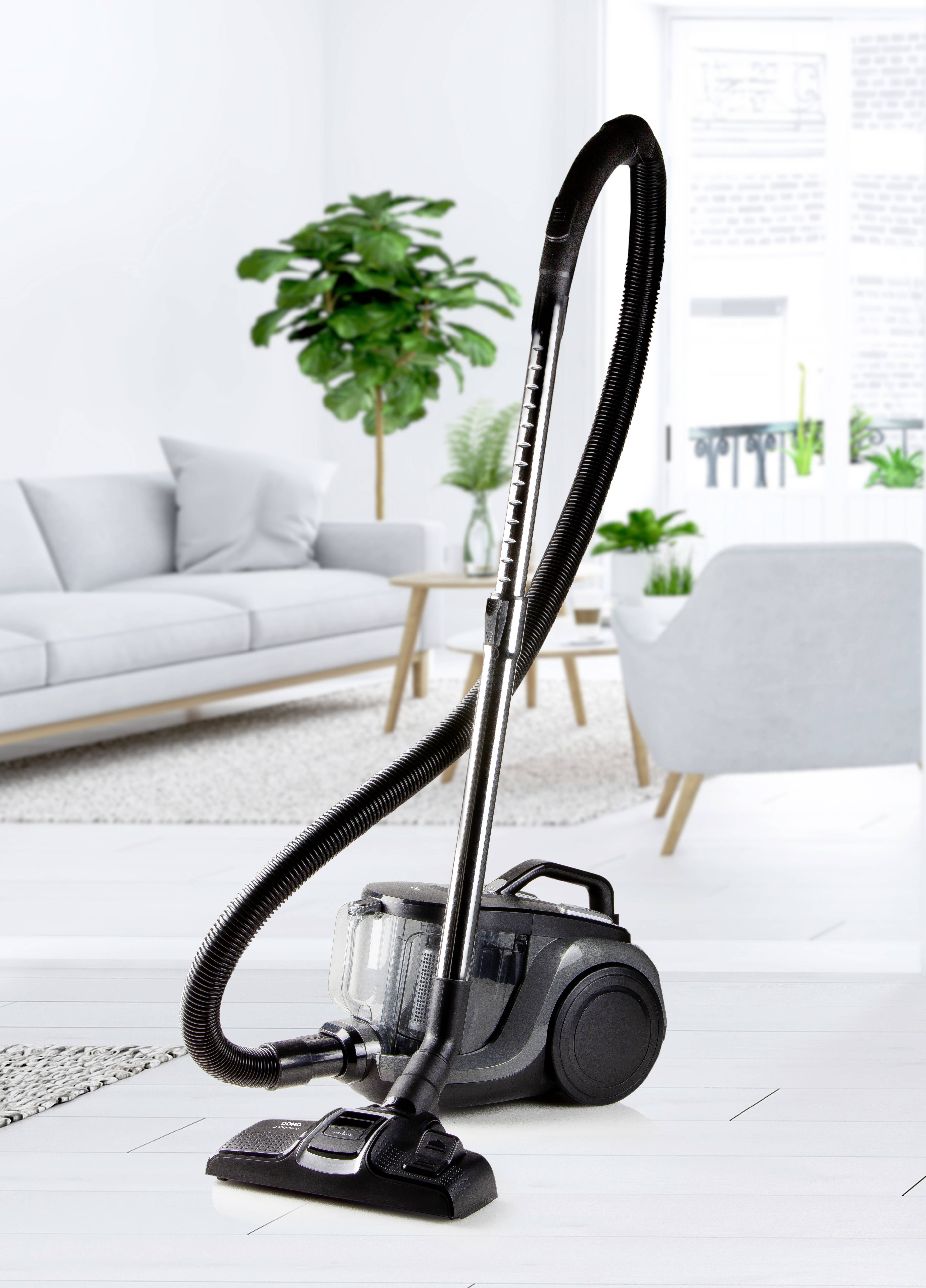 L'aspirateur est posé dans le salon sur un sol blanc, près d'un canapé gris et d'une plante verte, sous la lumière du jour provenant de grandes fenêtres.