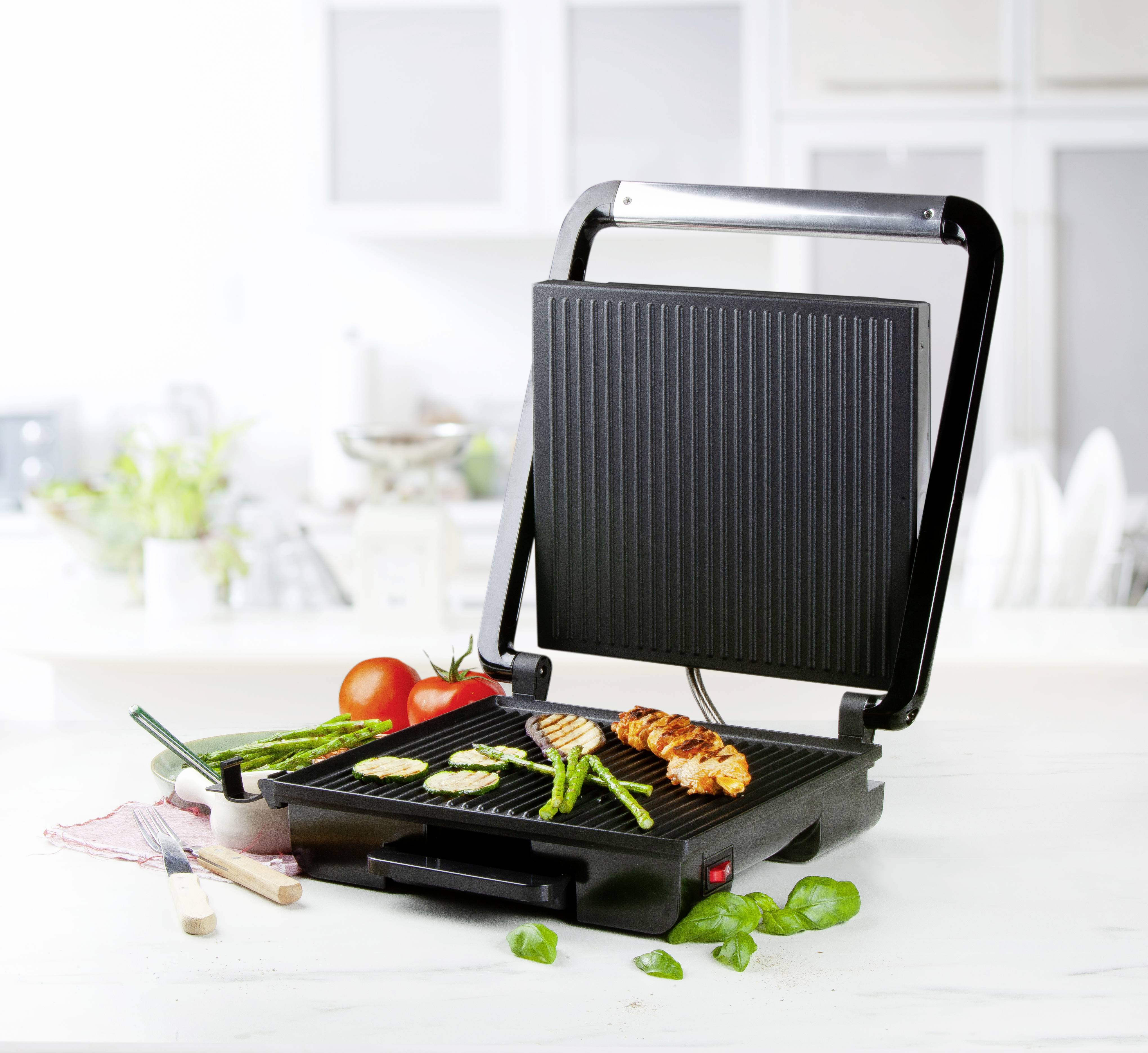 Grill électrique sur une table de cuisine avec des légumes et du poulet grillés. Des ustensiles de cuisine sont disposés en arrière-plan.