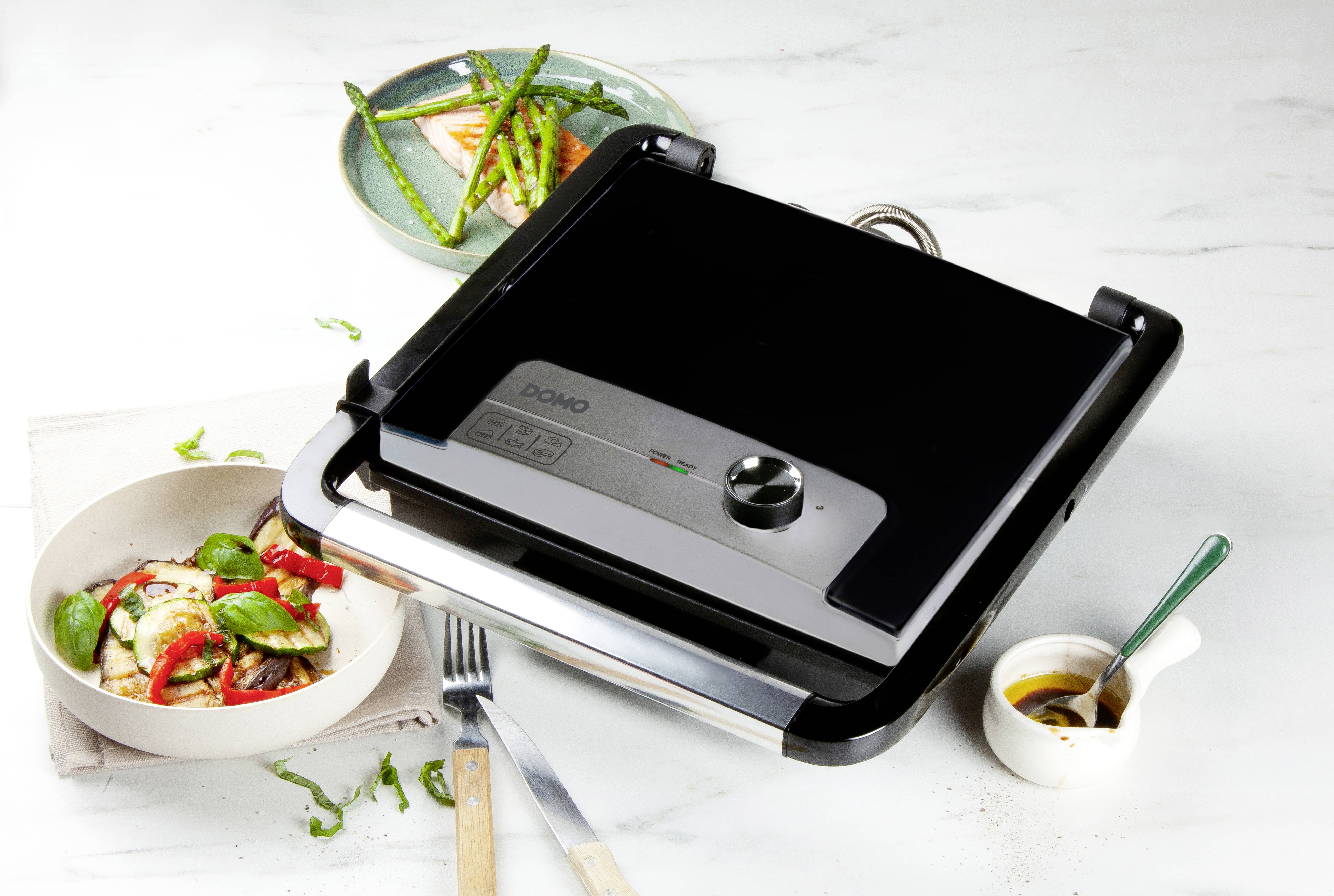 Un grill de table électrique posé sur une table à côté d'une assiette de légumes grillés, d'une salade et d'un petit bol de sauce.