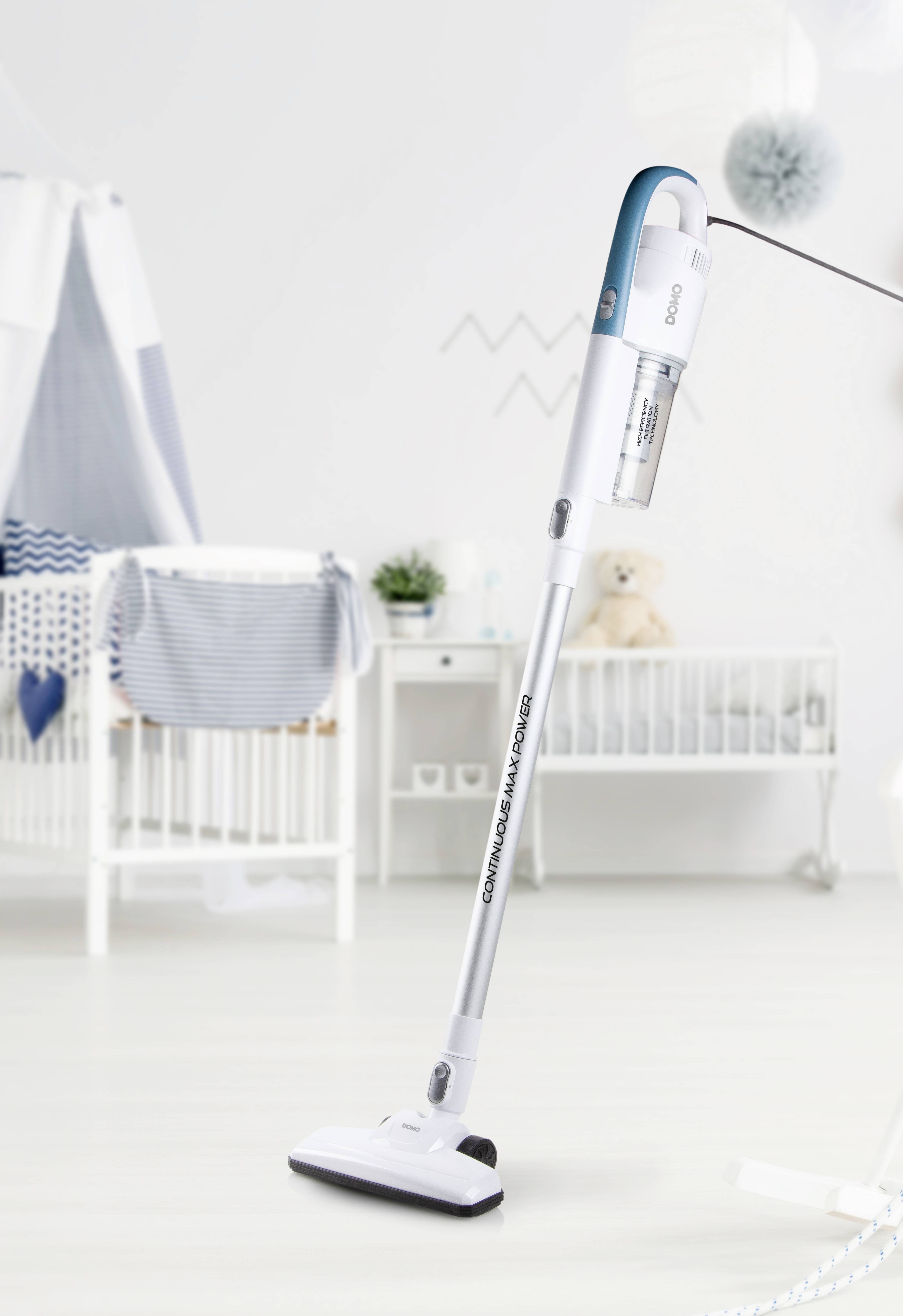 Un aspirateur sans fil est debout sur un sol clair dans une chambre d'enfant avec un berceau oscillant et des décorations discrètes.