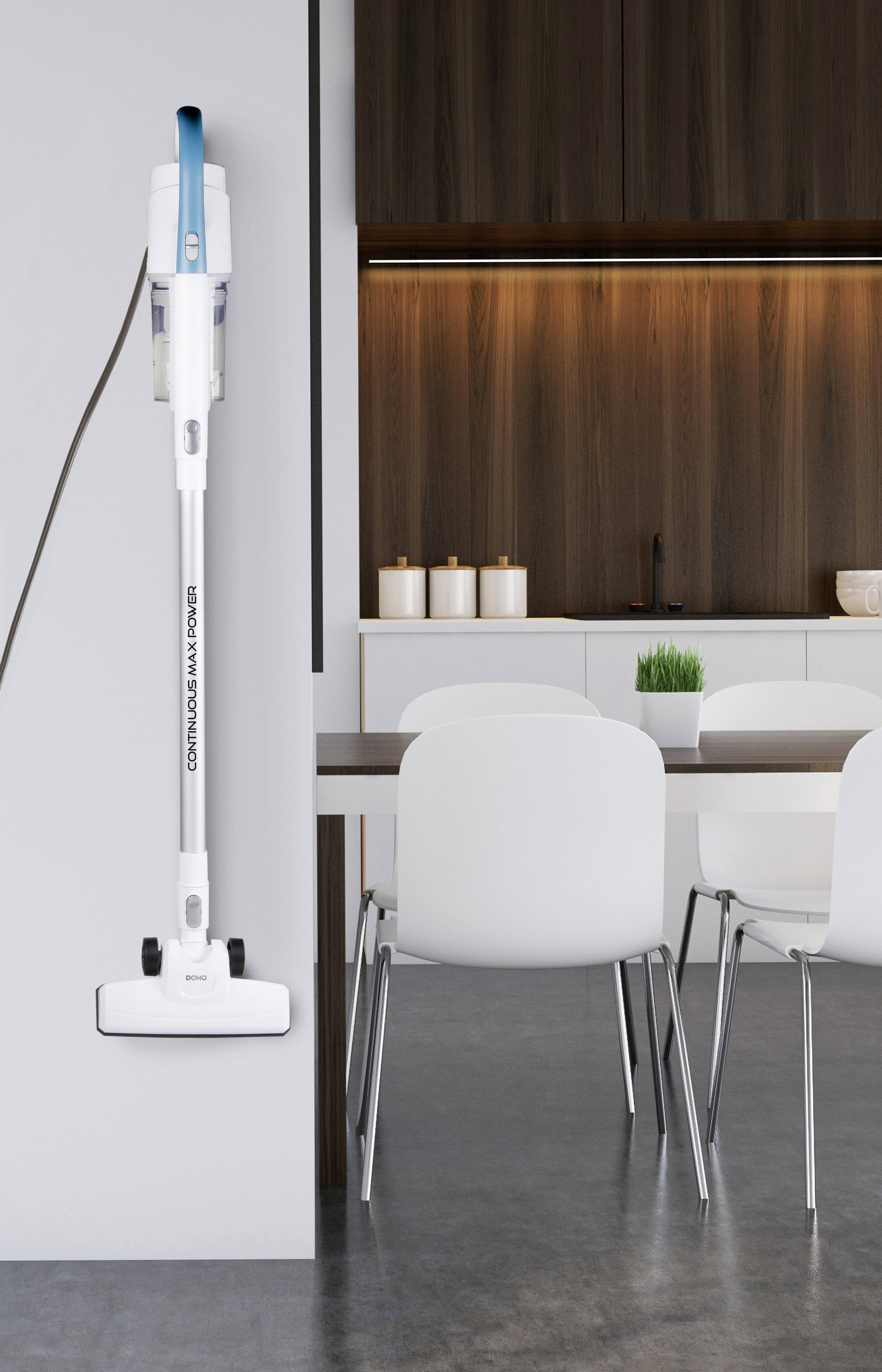 Un aspirateur blanc est suspendu à un mur clair dans une cuisine moderne avec des armoires en bois, des chaises blanches et une table.