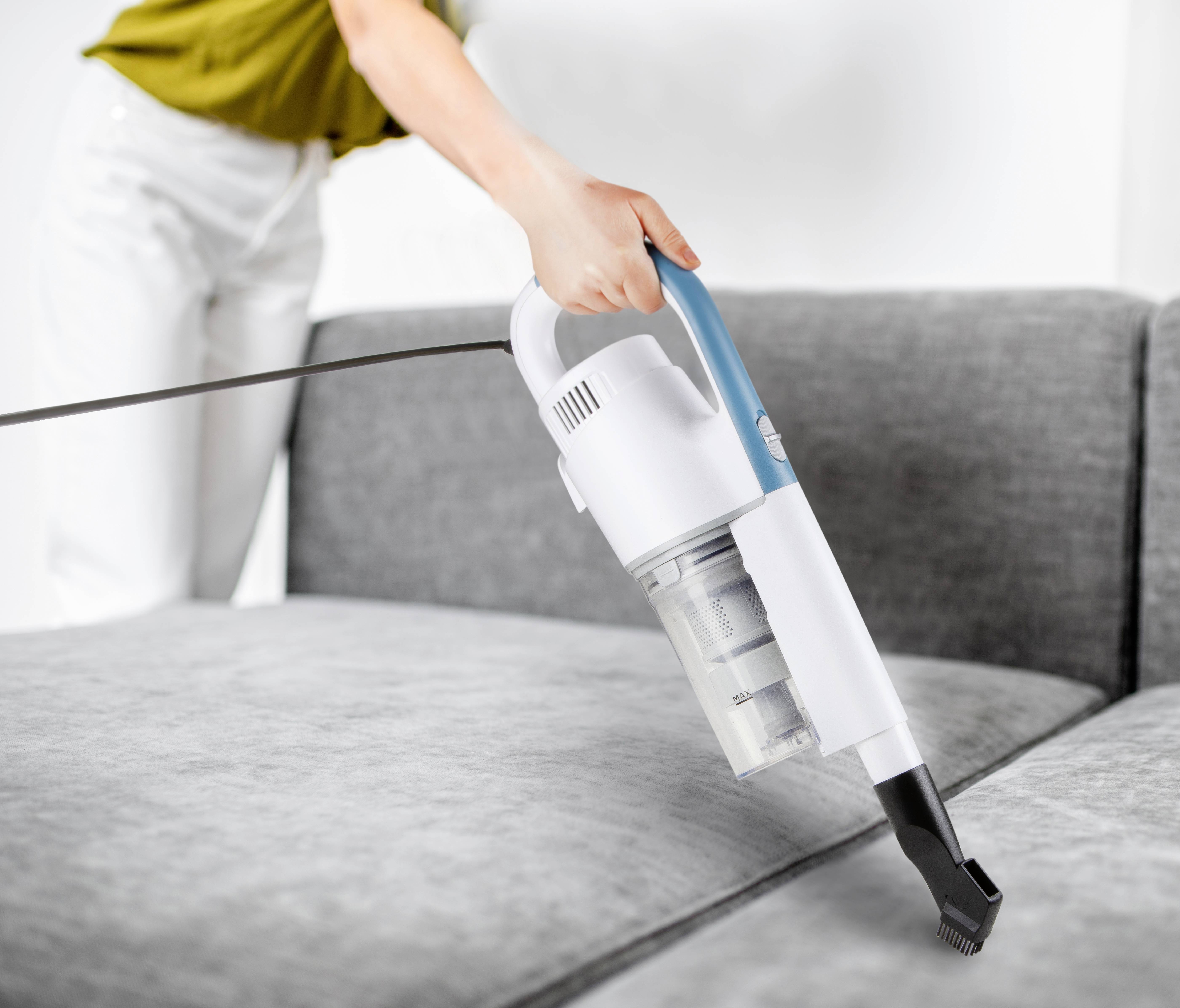 Une personne nettoie un canapé gris avec un aspirateur à main blanc. La personne porte des vêtements clairs.
