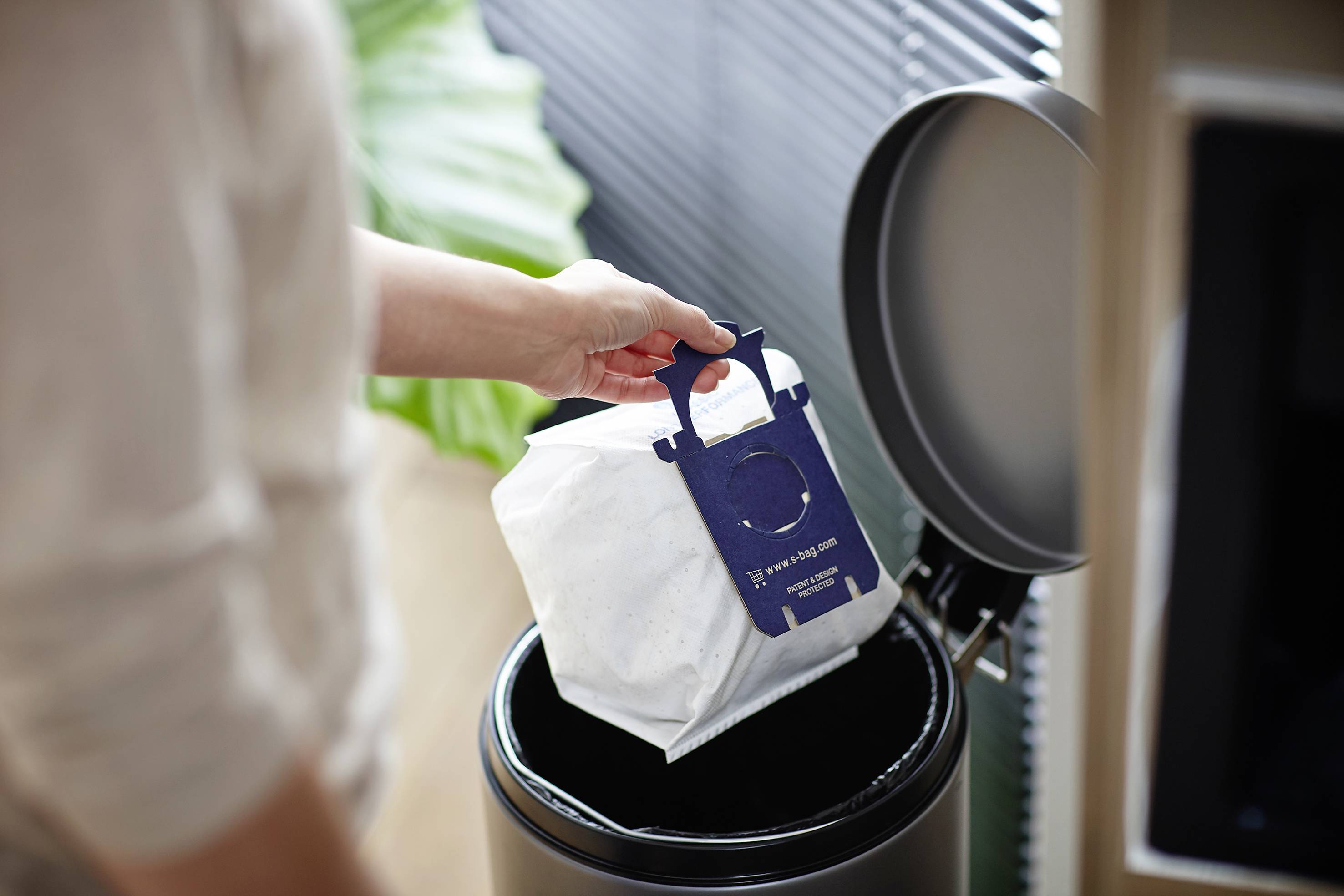 Une personne jette un sac d'aspirateur dans une poubelle. Une plante verte et des stores vénitiens sont visibles en arrière-plan.
