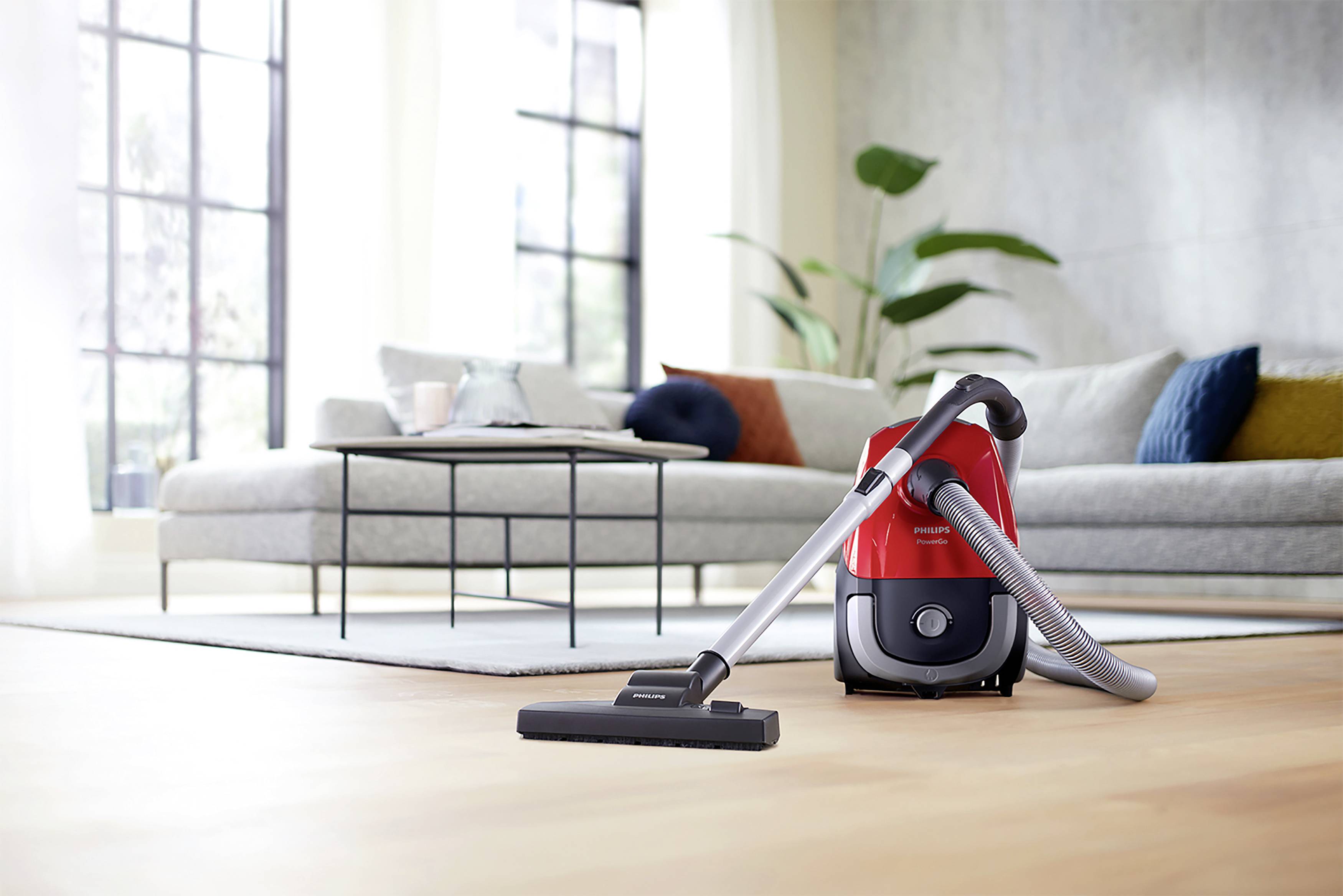 Un aspirateur rouge est posé sur un sol en bois clair devant un salon moderne avec un canapé, une table basse et de grandes fenêtres.