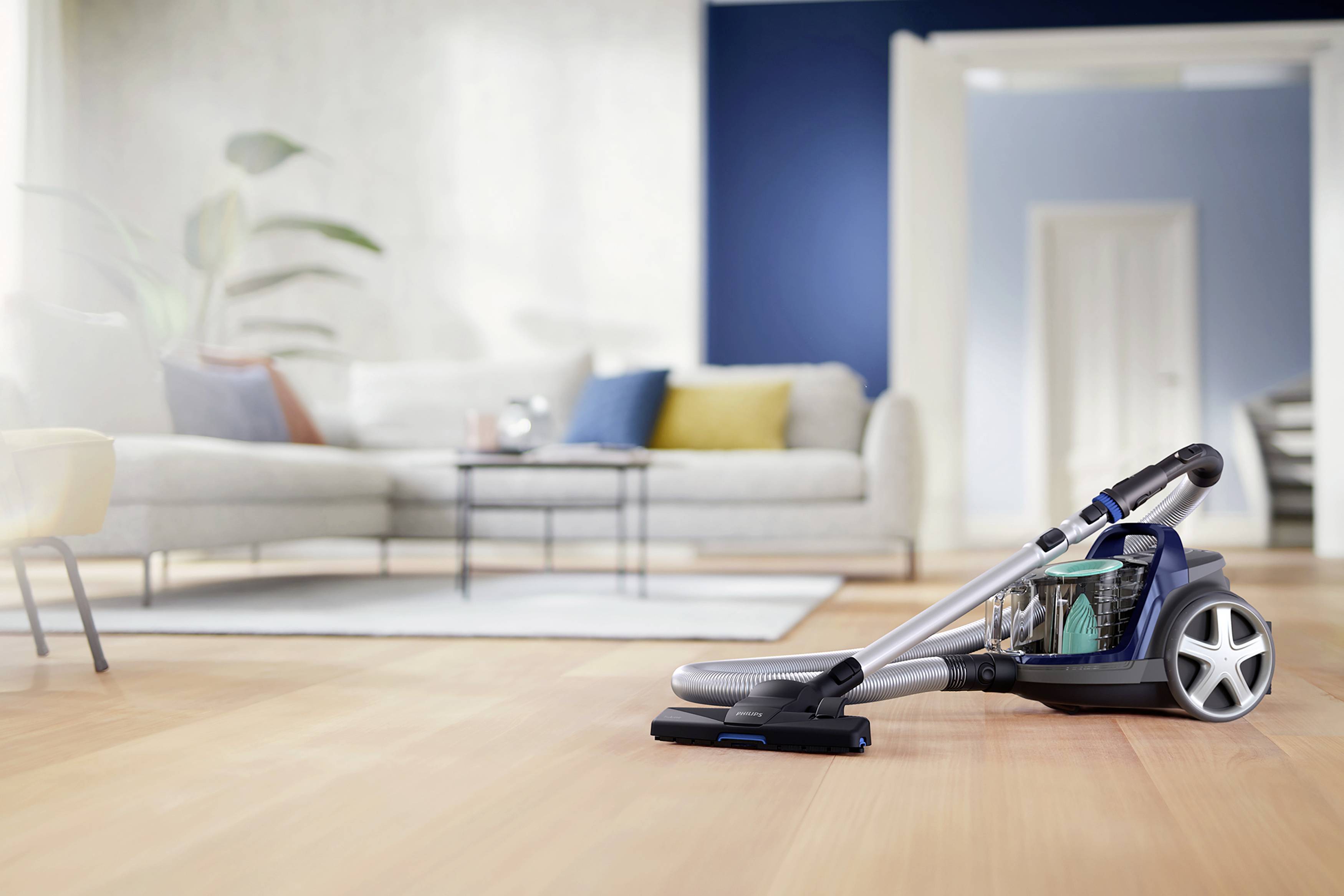 Un aspirateur sur un plancher en bois dans un salon au design moderne avec un canapé, des coussins et une petite table en arrière-plan.