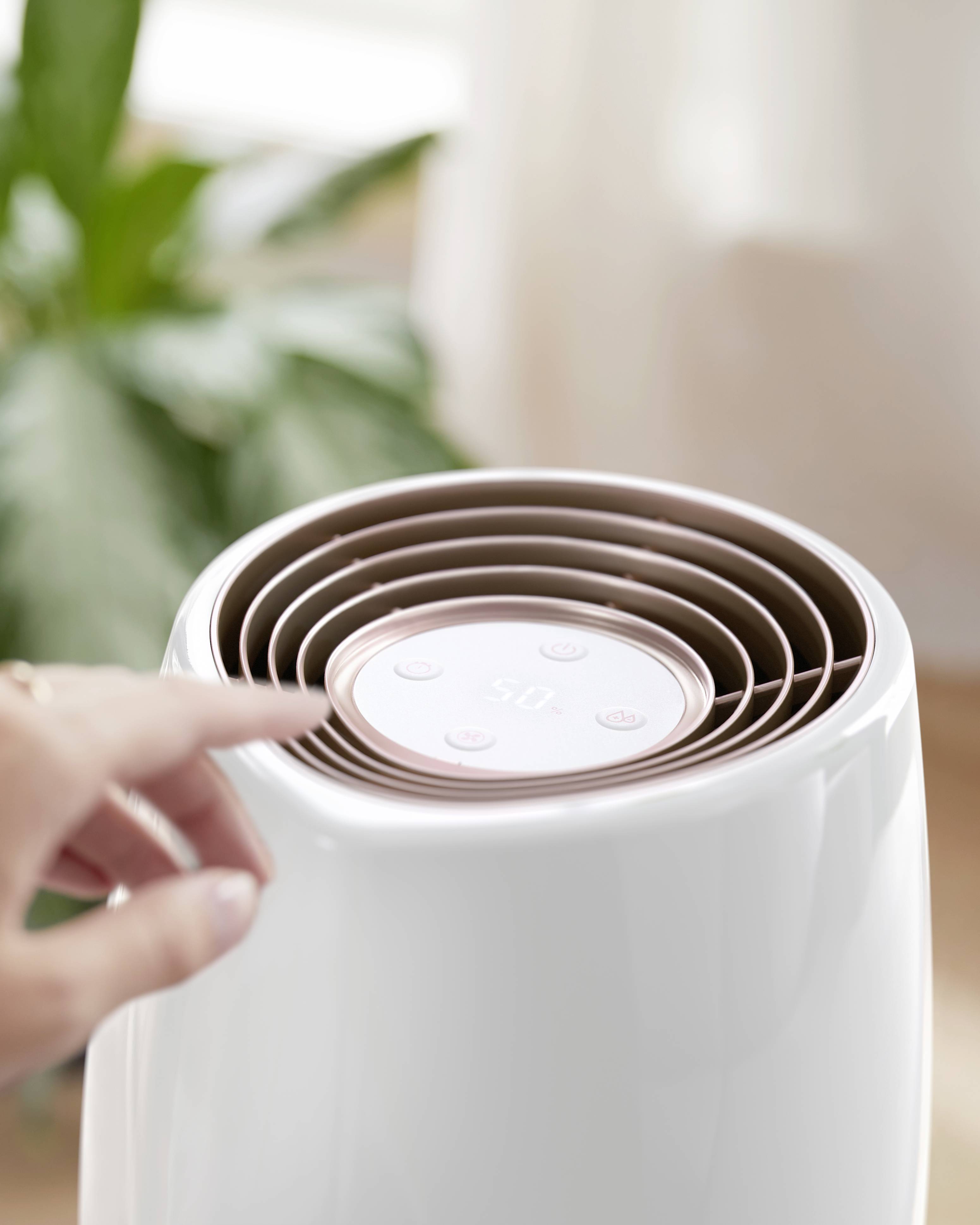 Un doigt touche les surfaces de commande supérieures d'un humidificateur. L'humidificateur est blanc avec des détails bruns. En arrière-plan, se trouve une plante verte.