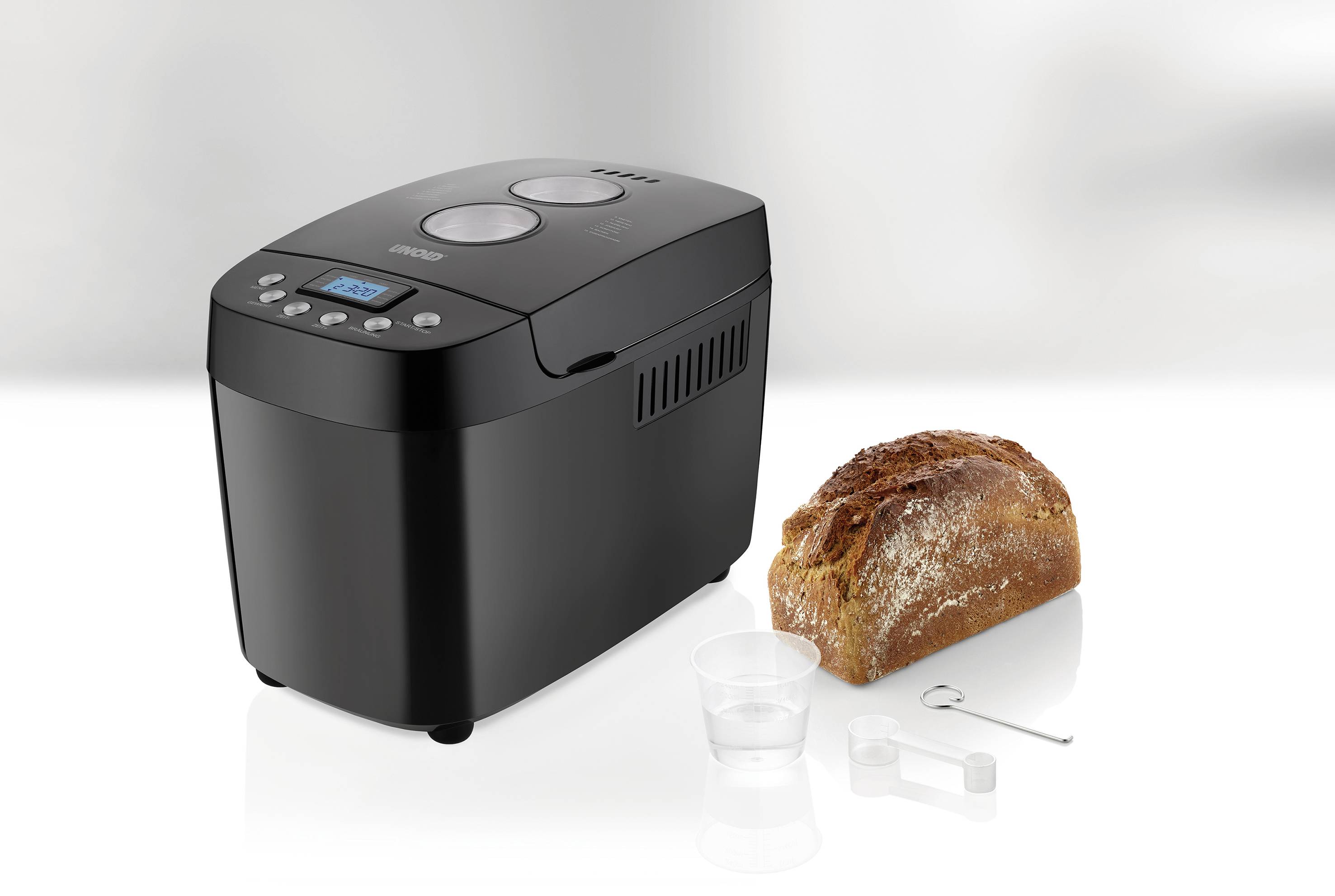 Machine à pain noire sur une cuisine avec un pain fraîchement cuit à côté. Accessoires comprenant un gobelet doseur et un crochet.