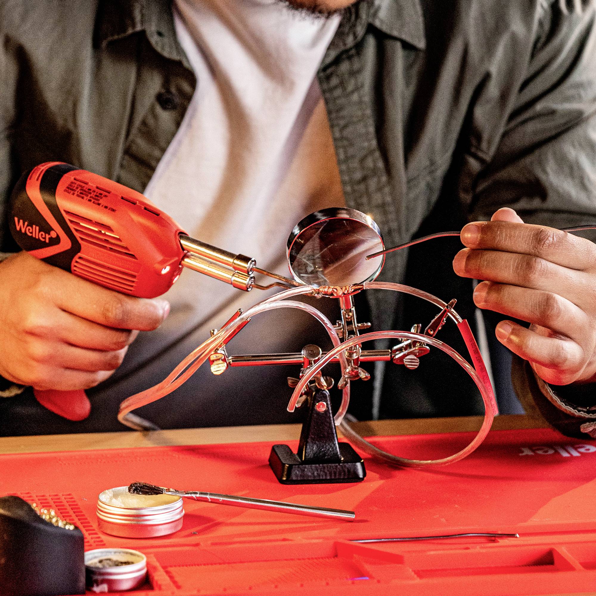 Une personne soude à la loupe et au fer à souder sur une structure métallique précise. D'autres outils sont posés sur la table.