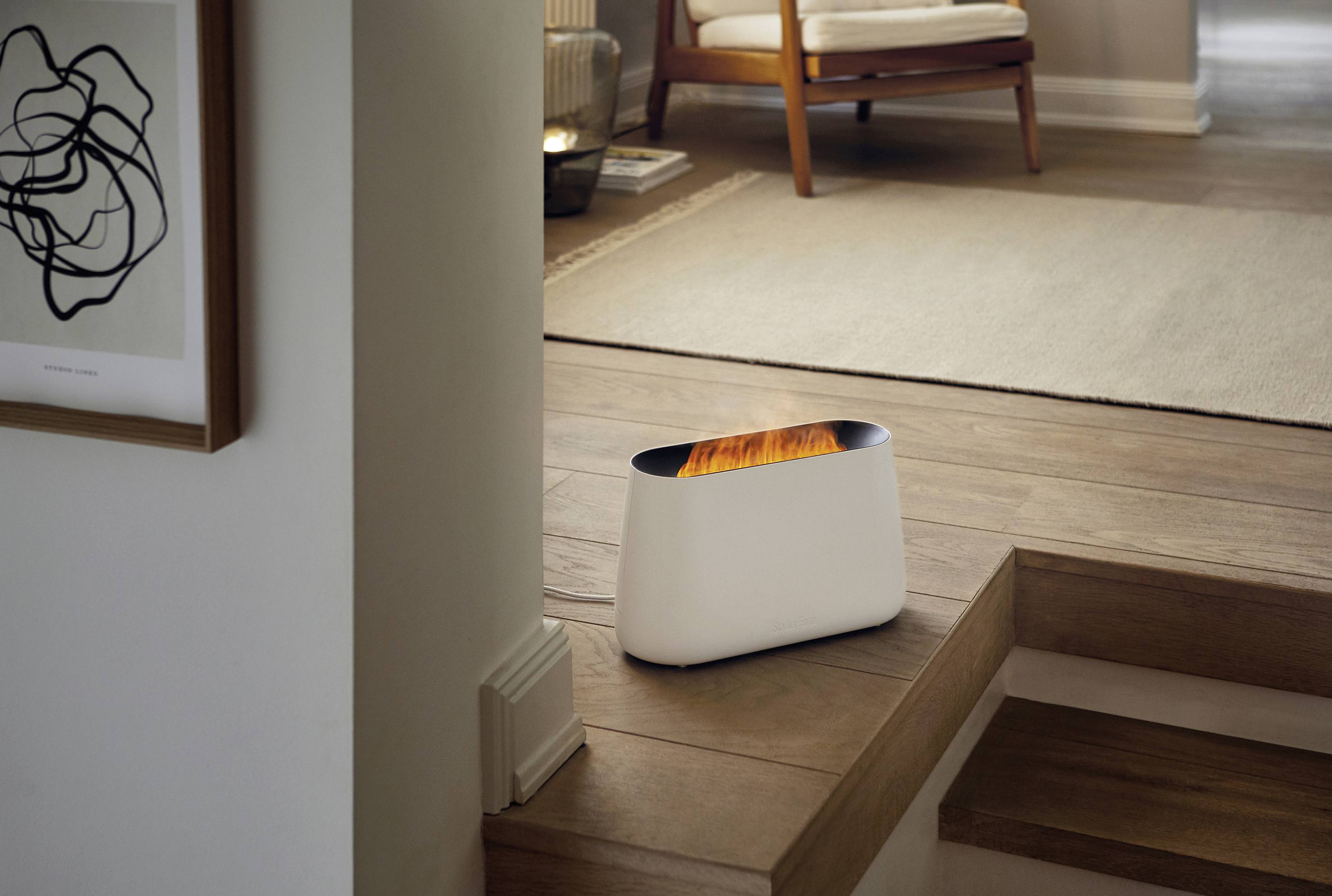 Un petit chauffage blanc se trouve dans un salon moderne à côté d'un escalier. À l'arrière-plan, on aperçoit un parquet et un fauteuil.