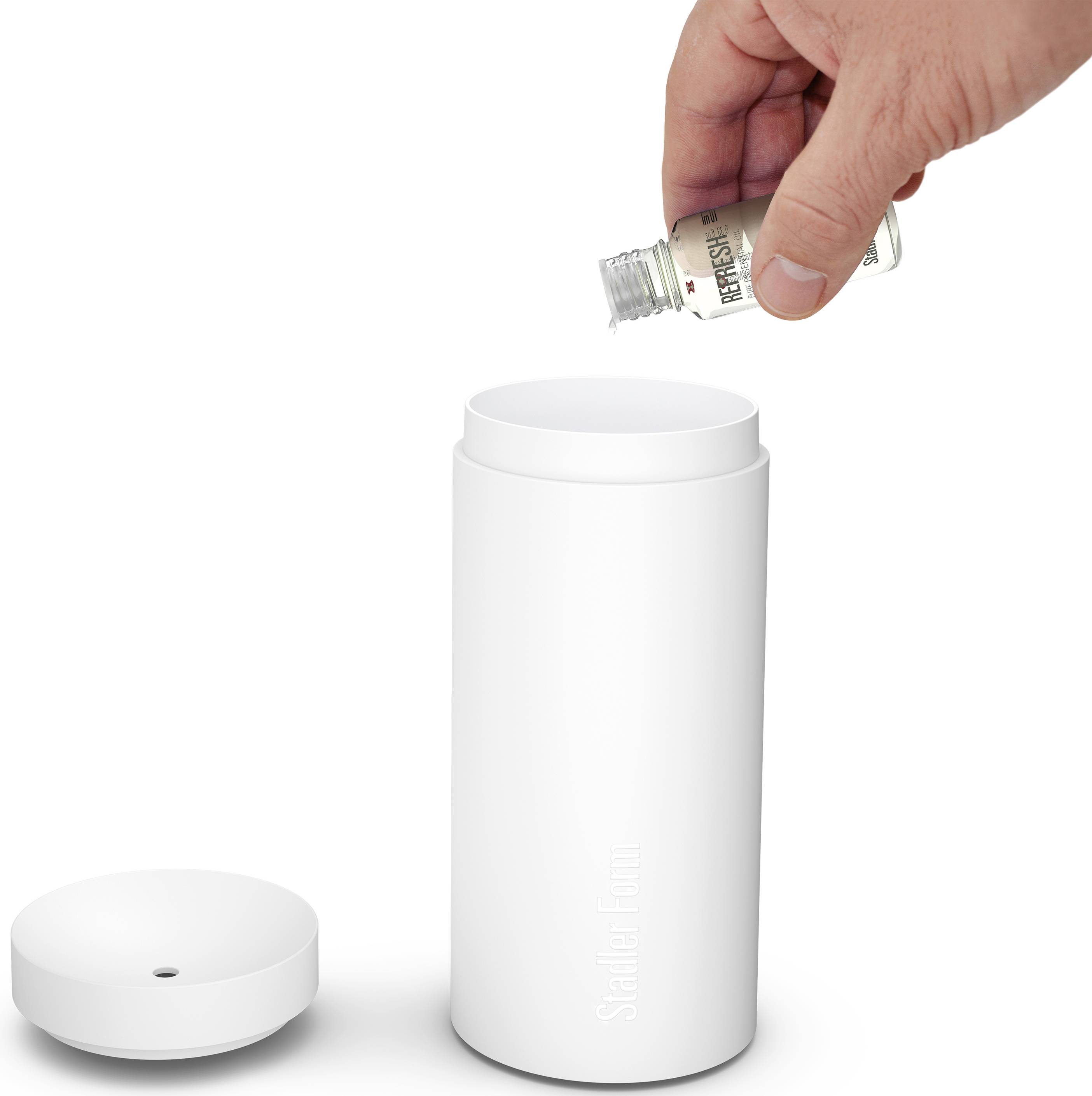 Une main verse de l'huile essentielle d'une petite bouteille dans un diffuseur d'aromathérapie blanc. Le couvercle est posé à côté.