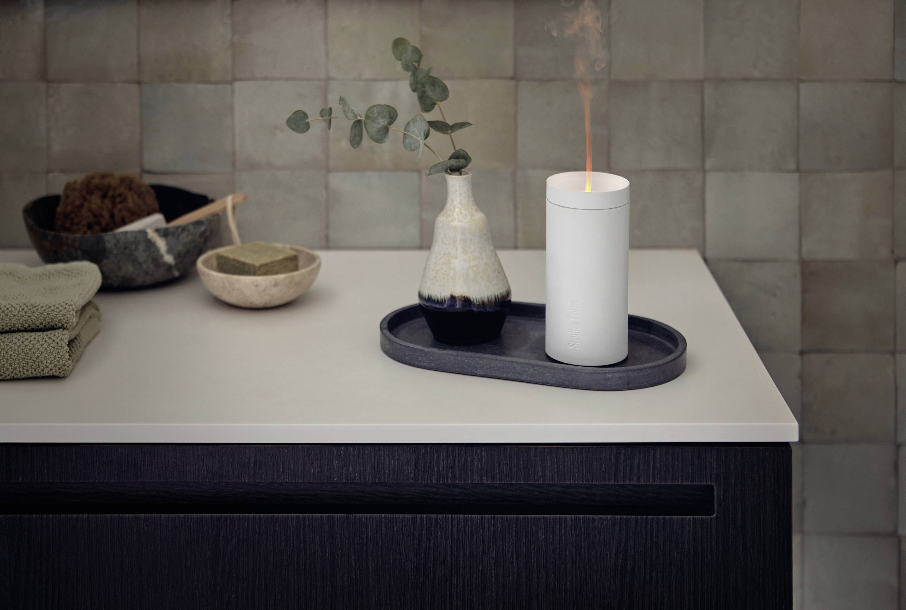 Une bougie brûle sur un plateau à côté d'un vase contenant une branche d'eucalyptus. Les deux sont posés sur un plan de travail moderne dans la salle de bain.