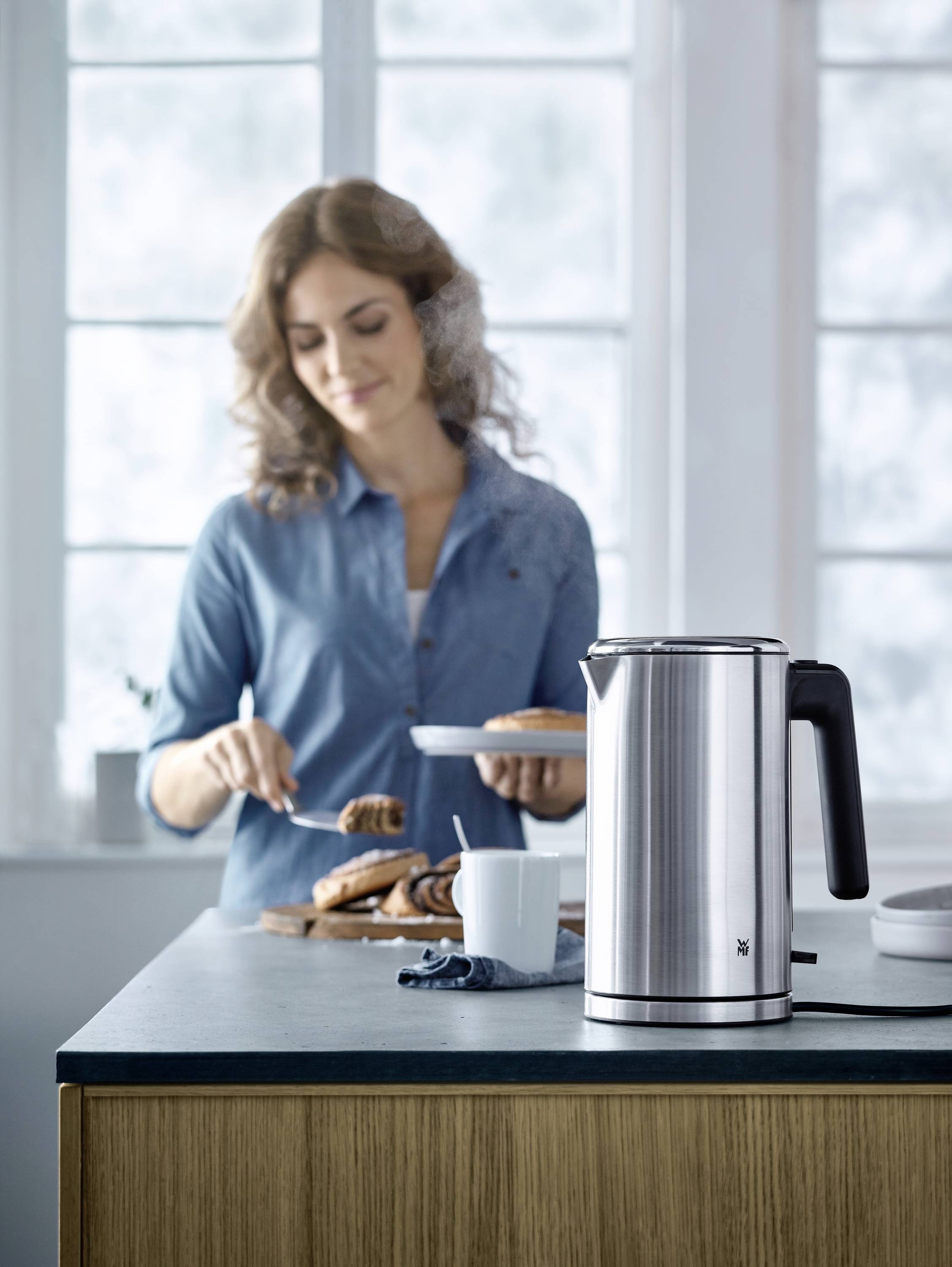 Une femme en arrière-plan dispose des pâtisseries sur une assiette. Au premier plan, une bouilloire en acier inoxydable est posée sur le comptoir de la cuisine.