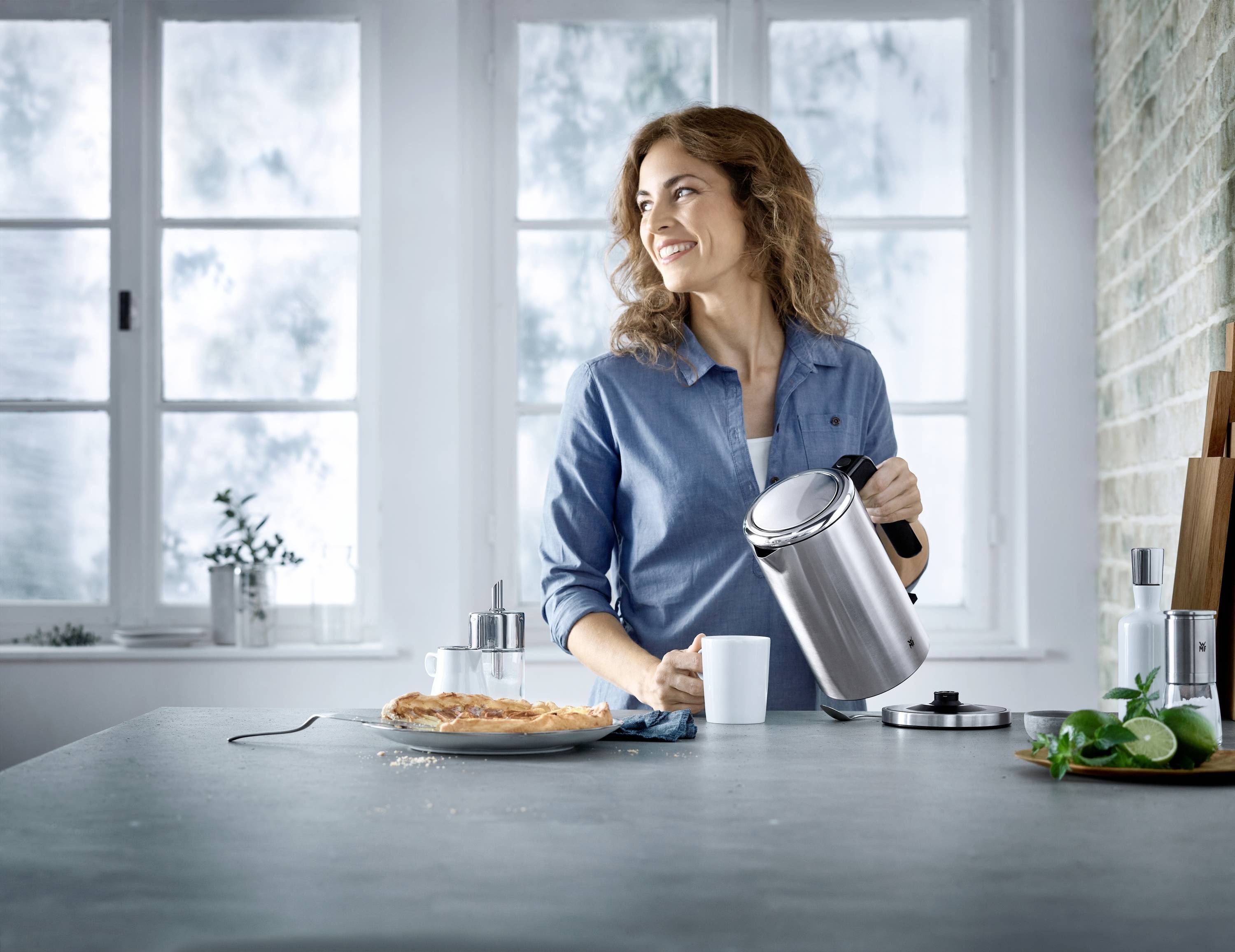 Une femme portant une chemise bleue verse de l'eau d'une bouilloire dans une tasse. Un gâteau et des ustensiles de cuisine sont posés sur la table.