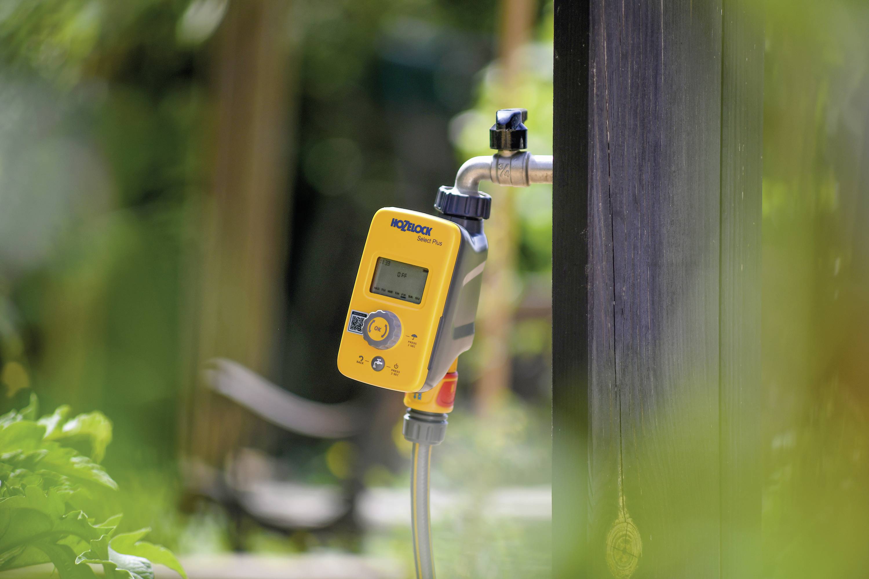 Un dispositif de commande d'irrigation jaune est connecté à un robinet dans le jardin. L'appareil est équipé d'un écran et de commandes.