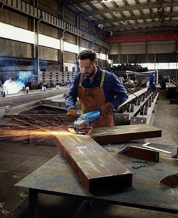 Un homme dans un atelier usine meule une pièce métallique avec une meuleuse d'angle, des étincelles volent. Des ouvriers sont en arrière-plan, dans un environnement industriel.