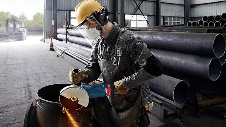 Un ouvrier en tenue de protection meule un tube avec une meuleuse d'angle dans un hall d'usine. En arrière-plan : des piles de tubes.