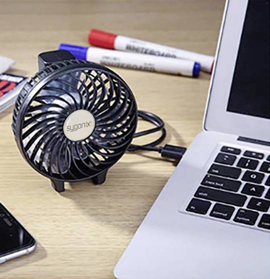 Un petit ventilateur de table noir avec connexion USB est posé à côté d'un ordinateur portable. À l'arrière-plan, deux stylos sont posés sur la table.