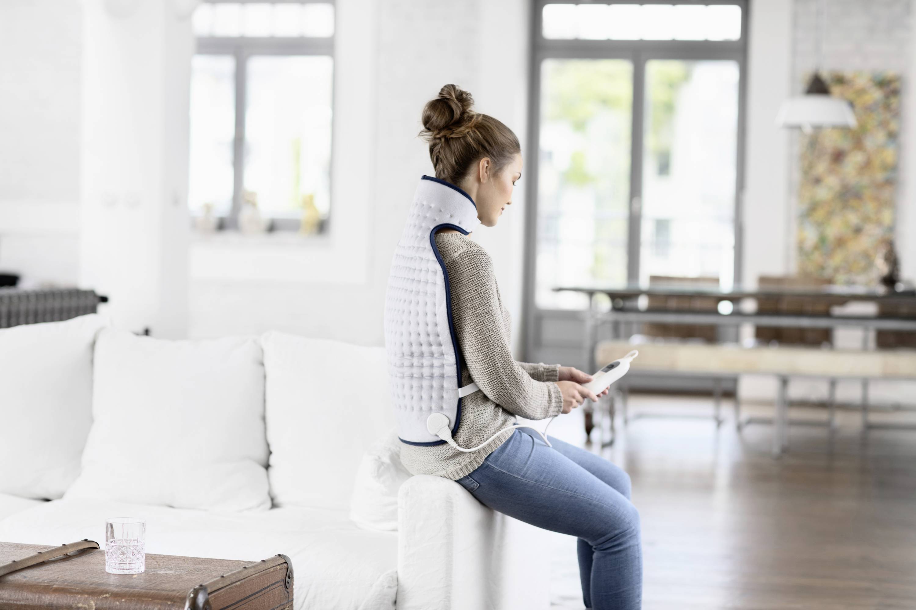Une femme est assise sur un canapé, portant un gilet chauffant à dos, et tient une tablette dans un salon lumineux.
