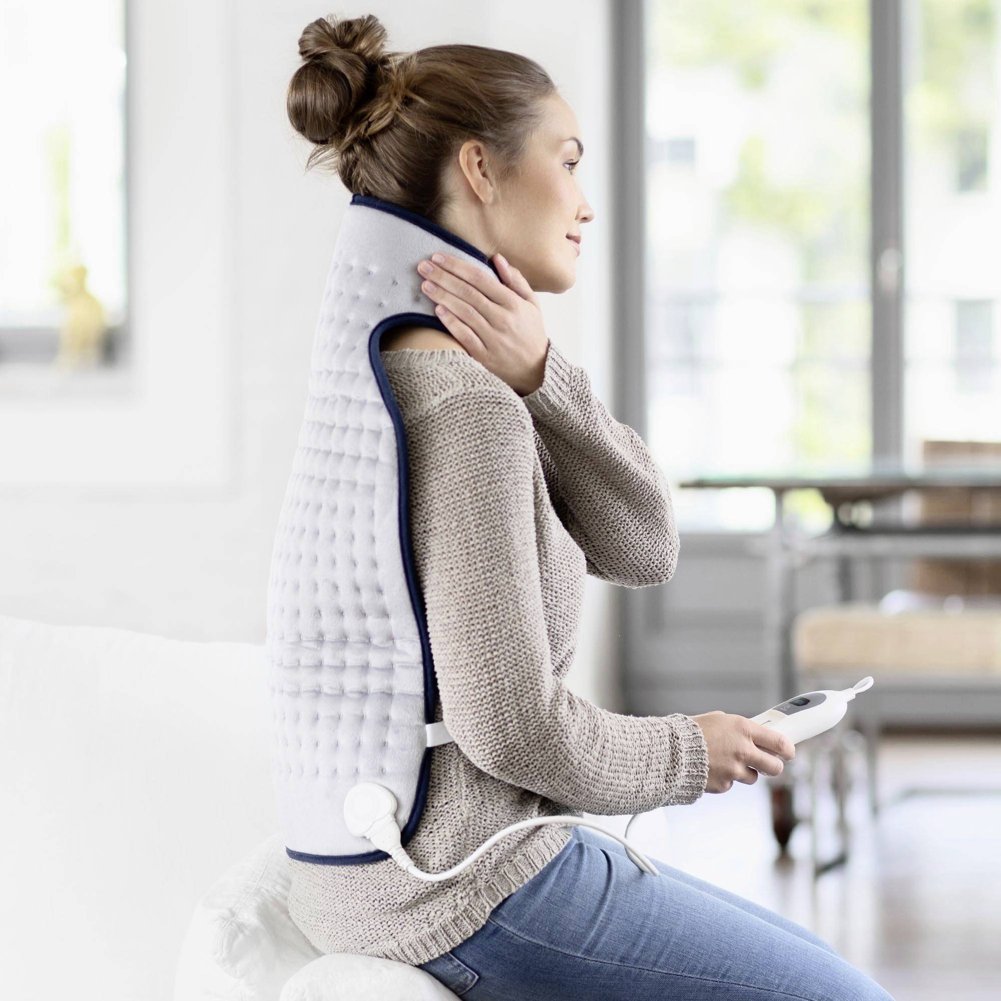 Une femme est assise sur un canapé et porte un coussin chauffant pour le dos. Elle tient la télécommande de l'appareil.