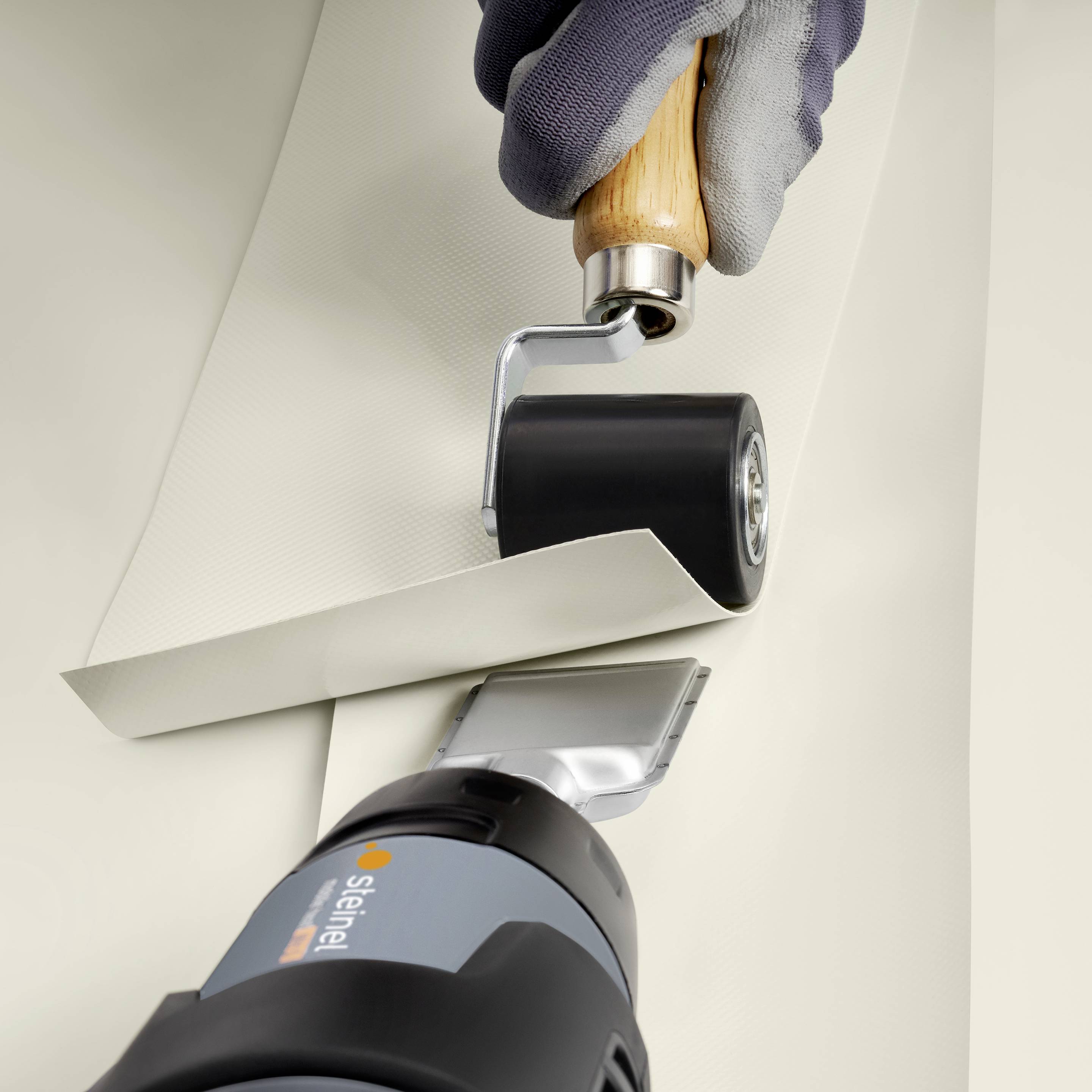 Une main gantée tient un outil qui relie une couture sur un matériau textile beige. Un autre manche se trouve en dessous.