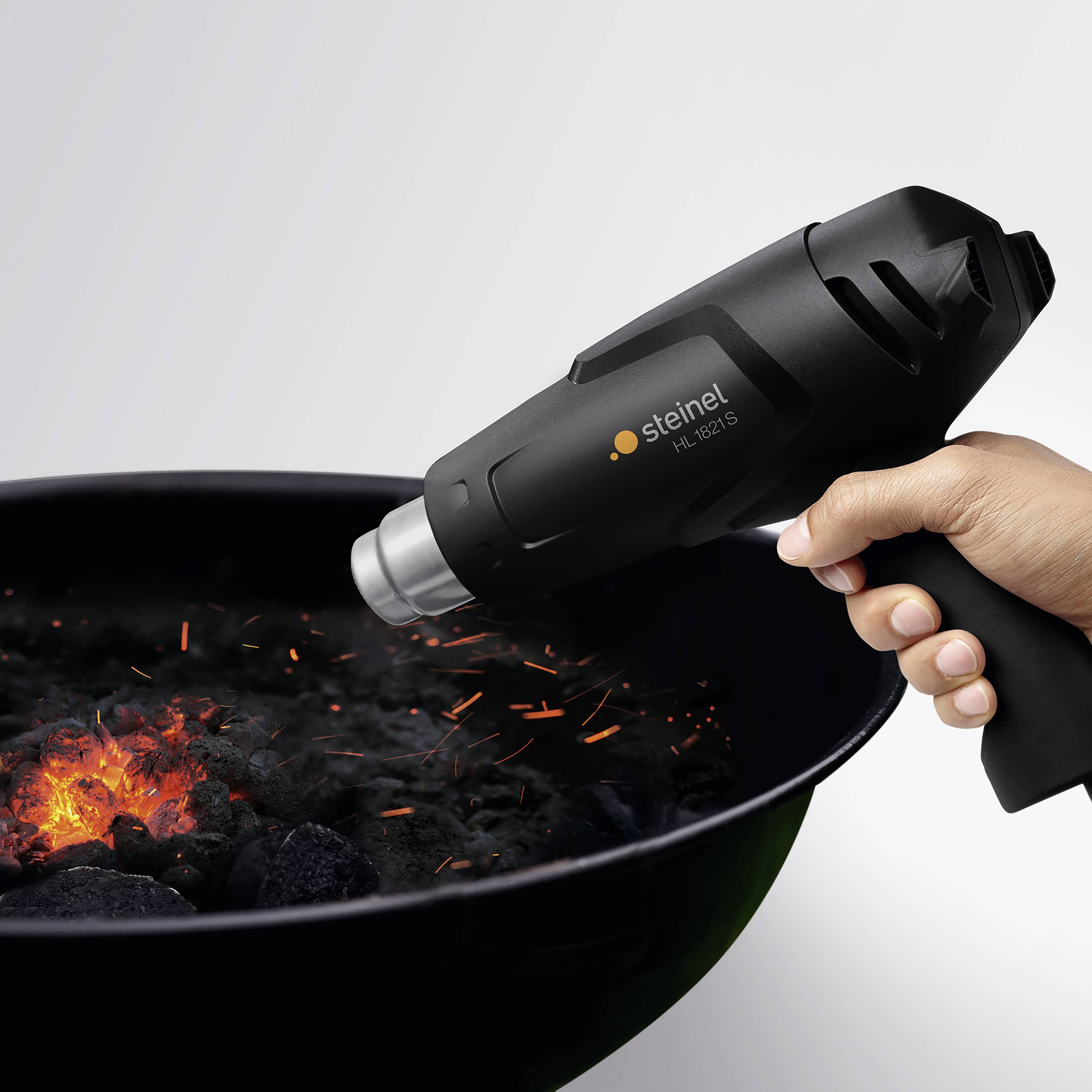 Une main tient un pistolet thermique au-dessus de briquettes de charbon incandescentes, des étincelles volent.
