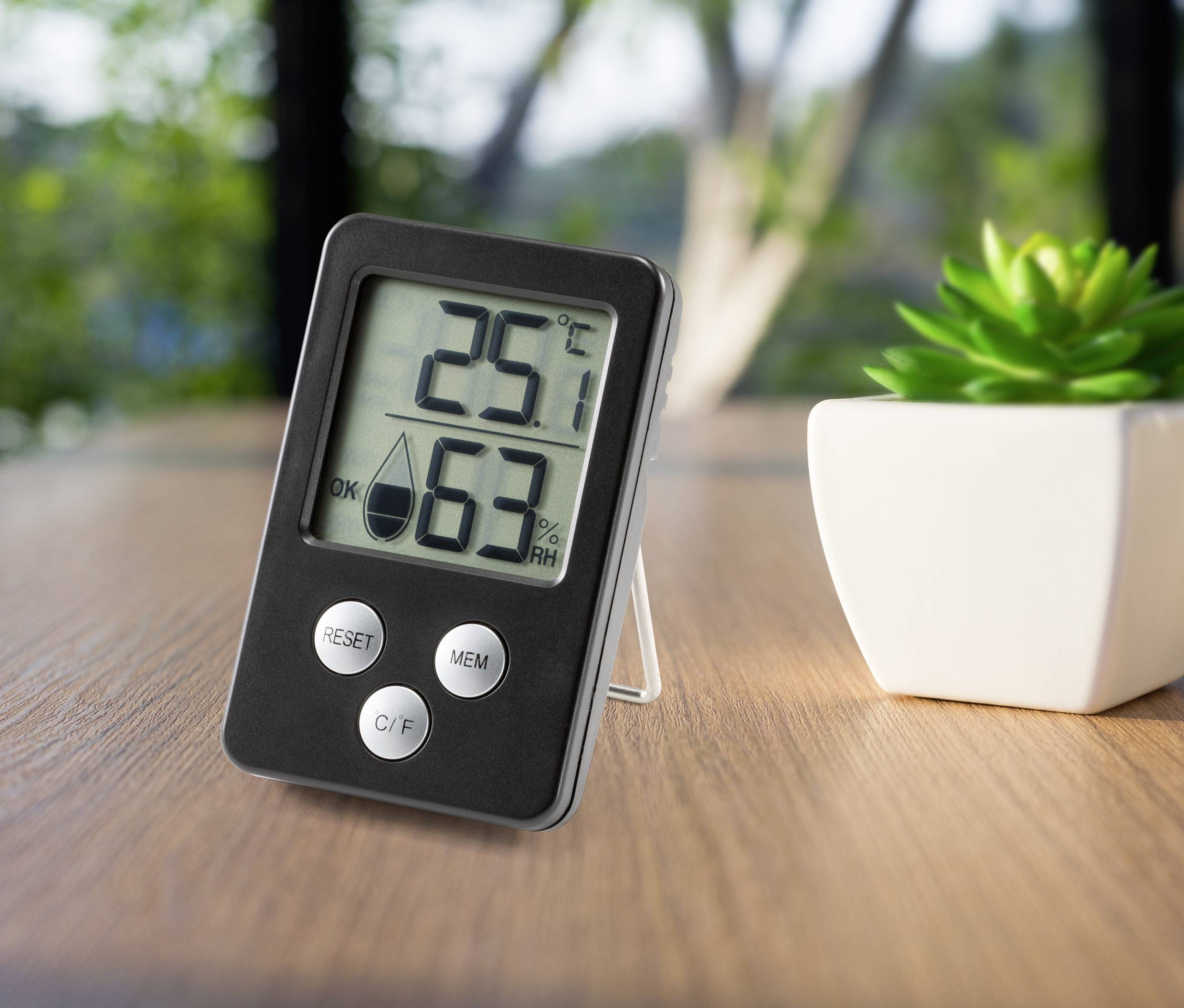 Thermomètre numérique et hygromètre posés sur une table, affichant 25°C et 63% d'humidité. Une plante est visible en arrière-plan.
