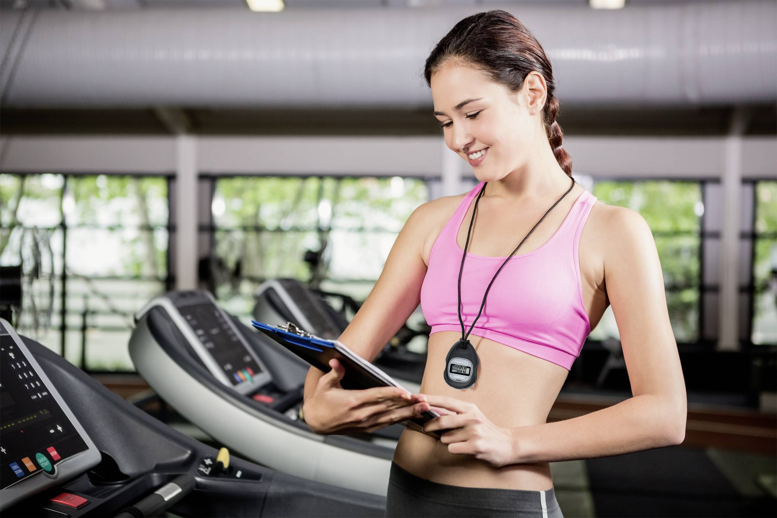 Une femme souriante se tient devant des tapis de course dans une salle de sport, tenant un presse-papiers et vérifiant des données sur un podomètre.