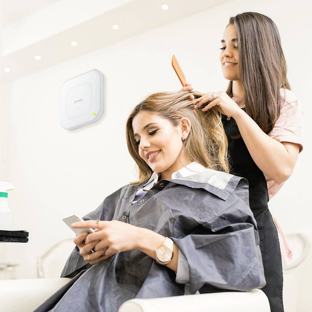 Deux femmes dans un salon de coiffure : Une est assise, portant une cape de coiffure, souriante et utilisant son téléphone portable ; l'autre lui coiffe les cheveux. Un routeur Wi-Fi est visible en arrière-plan.