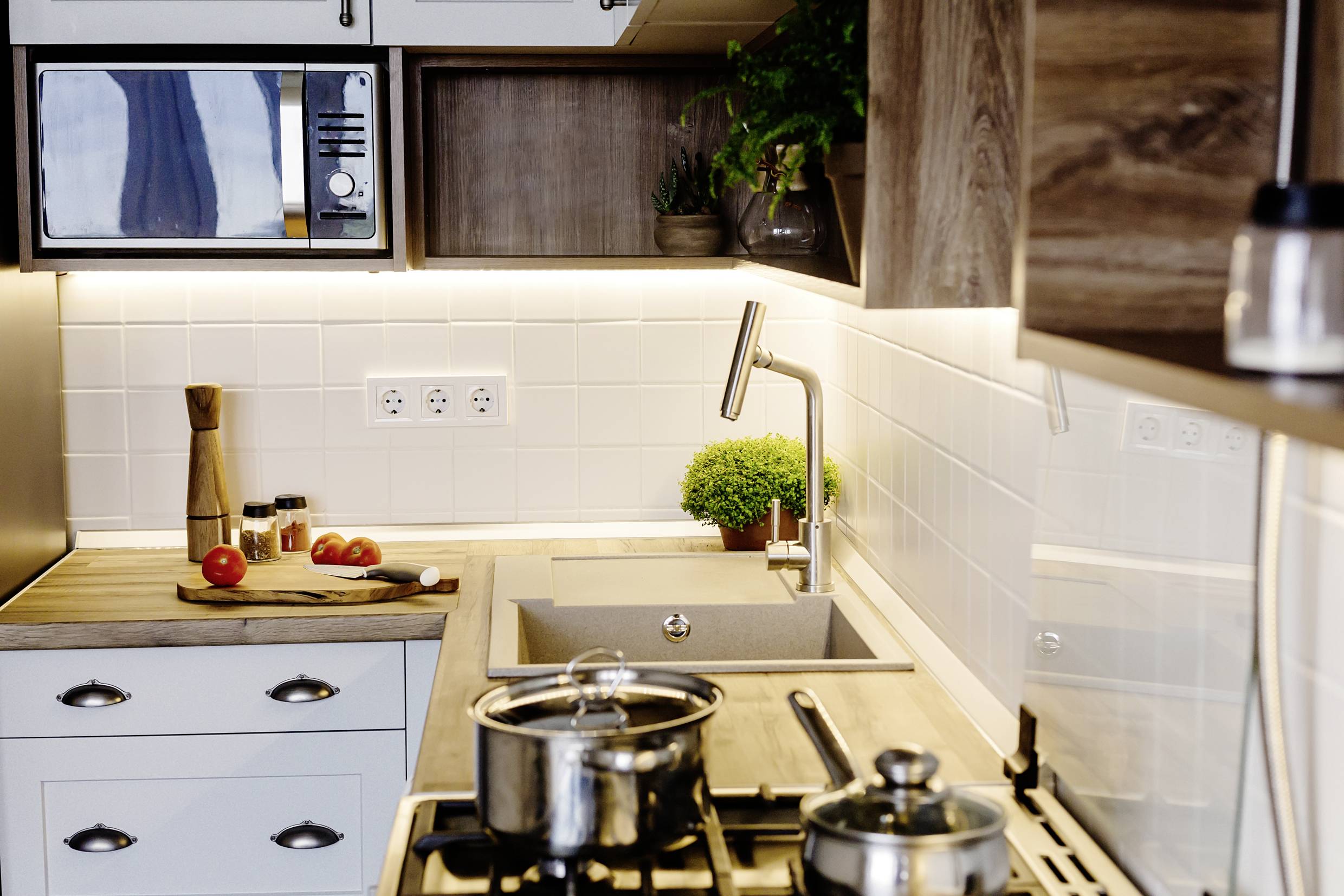 Cuisine moderne avec cuisinière, casseroles, évier, plantes et légumes sur le plan de travail ; carrelage blanc et meubles en bois.