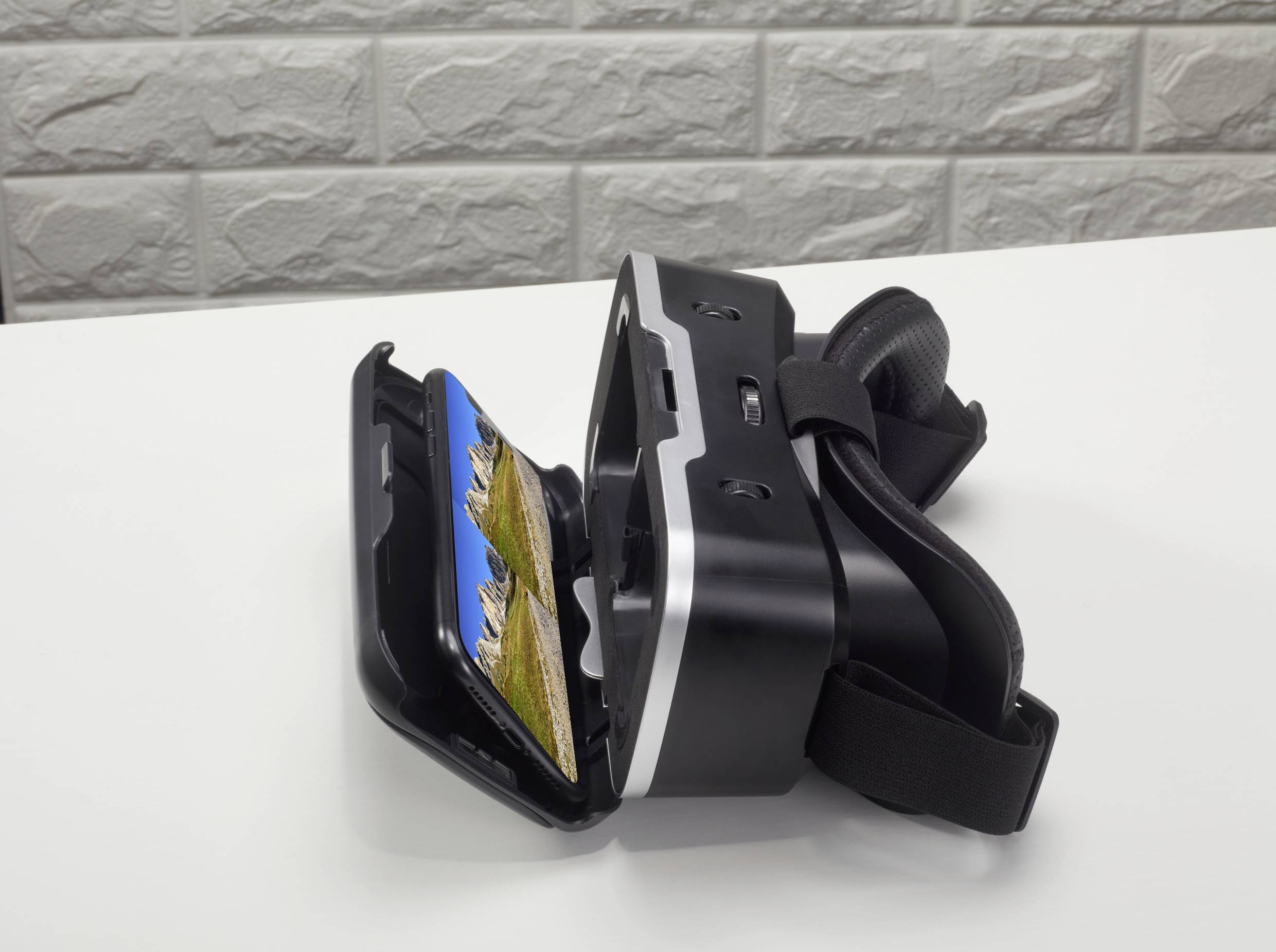 Casque de réalité virtuelle sur une table blanche, ouvert avec un smartphone affichant un paysage montagneux. Mur de briques en arrière-plan.