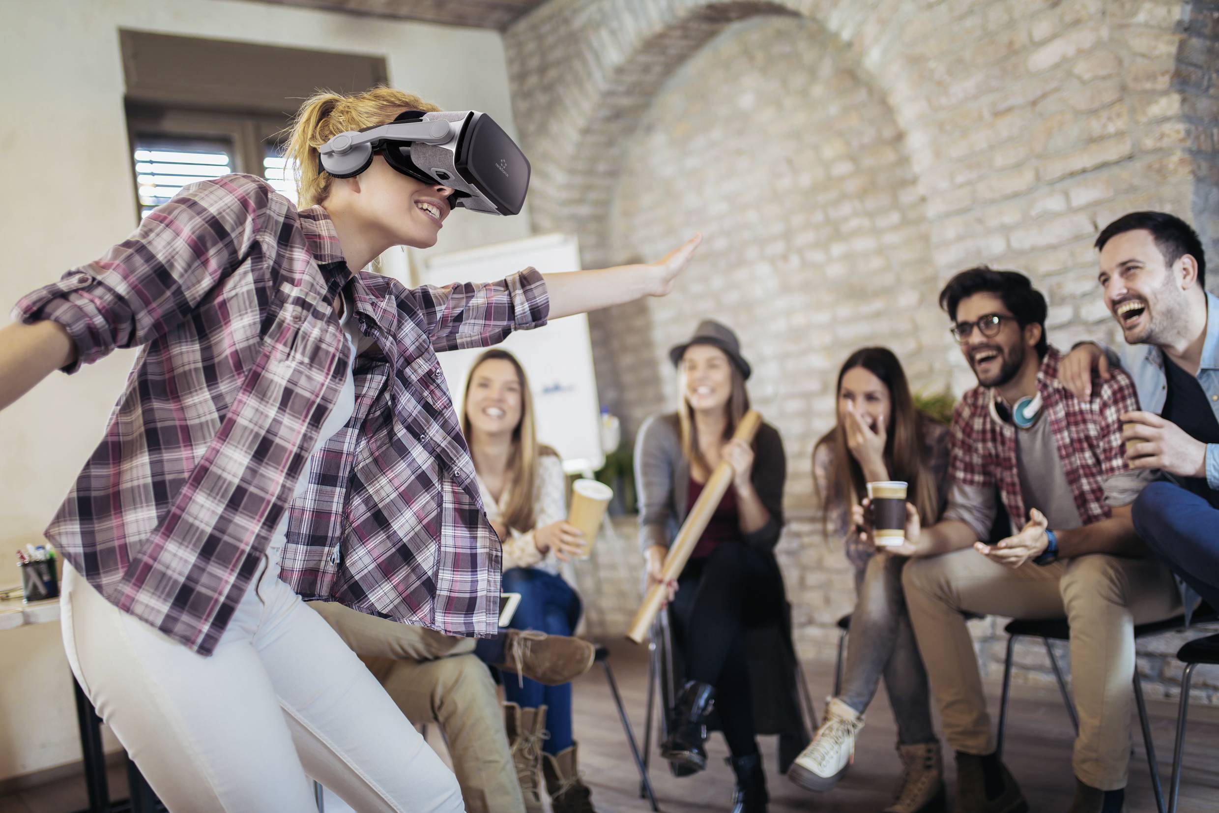 Une femme porte un casque de réalité virtuelle et bouge, tandis que cinq personnes rient en la regardant. Tous sont installés confortablement dans une pièce conviviale.