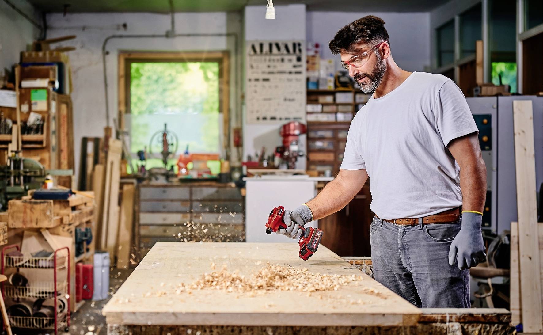 Un homme travaille le bois avec une machine rouge et noire dans un atelier, des copeaux de sciure volent. Des étagères et des fenêtres sont visibles en arrière-plan.