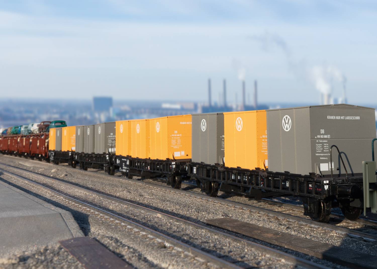 Plusieurs wagons de marchandises portant des conteneurs orange et gris de la marque VW sont garés sur une voie ferrée, avec des cheminées visibles en arrière-plan.