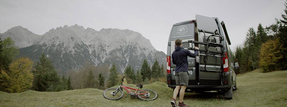 Une personne se tient à côté d'un fourgon sur un pré en montagne et range un vélo. Des arbres et une chaîne de montagnes sont visibles en arrière-plan.