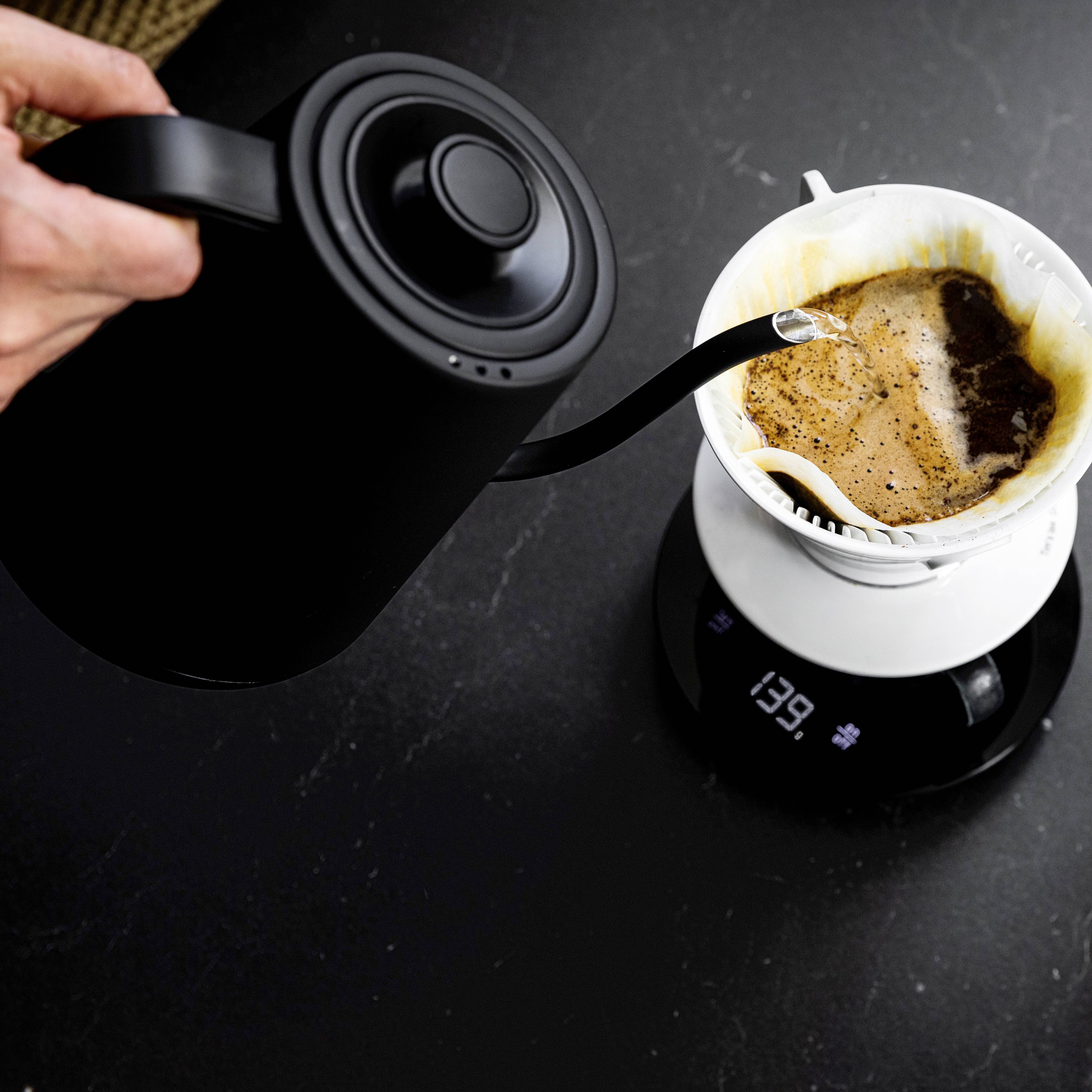 Une main verse de l'eau d'une bouilloire noire dans un filtre à café blanc posé sur une balance. De la vapeur s'élève.