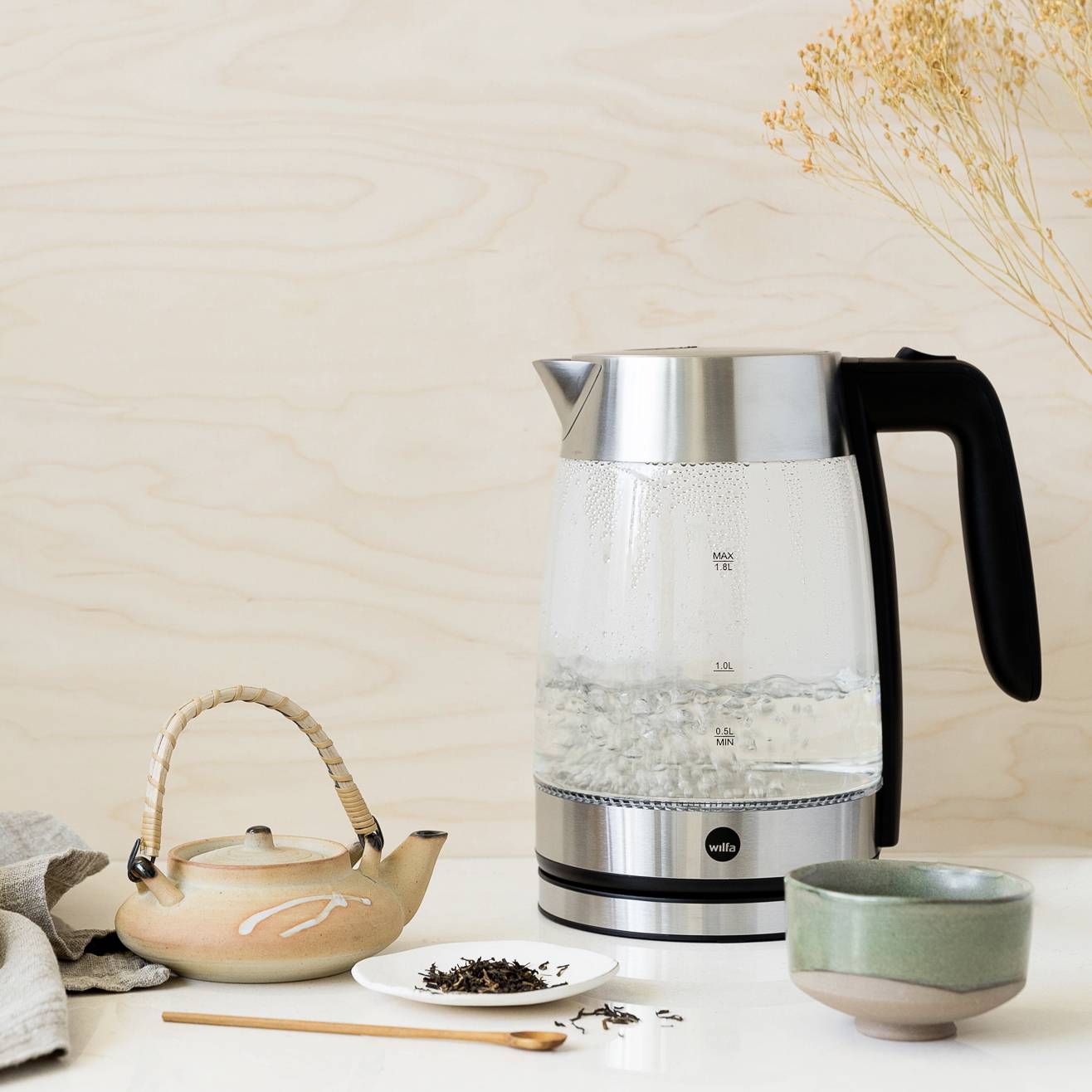 Une bouilloire en verre remplie d'eau bouillante est posée sur une table. À côté, une théière, une tasse et des feuilles de thé sur une cuillère.