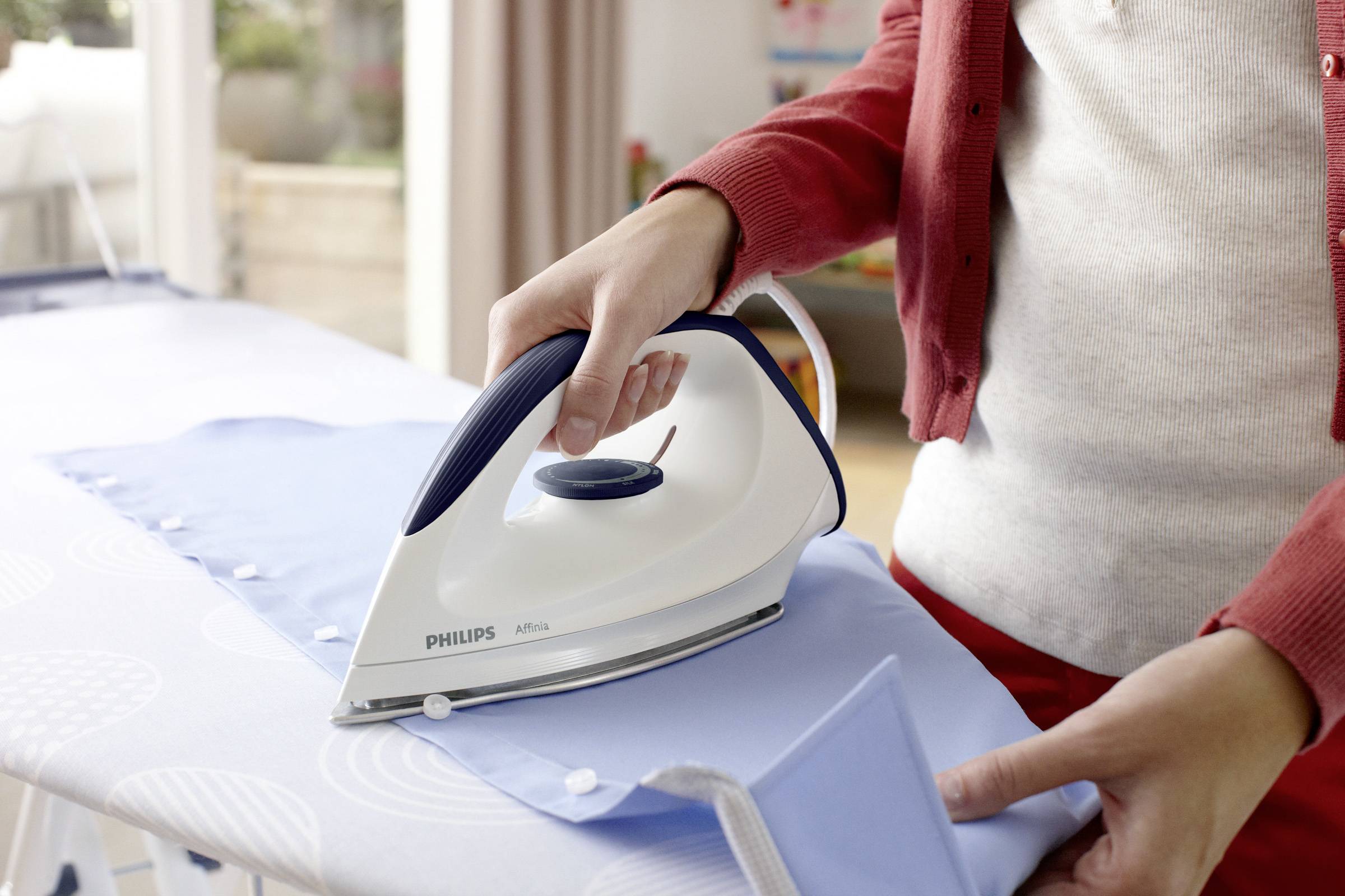 Une personne repasse une chemise bleu clair sur une planche à repasser. Le fer à repasser est de la marque Philips. La lumière entre par une fenêtre.