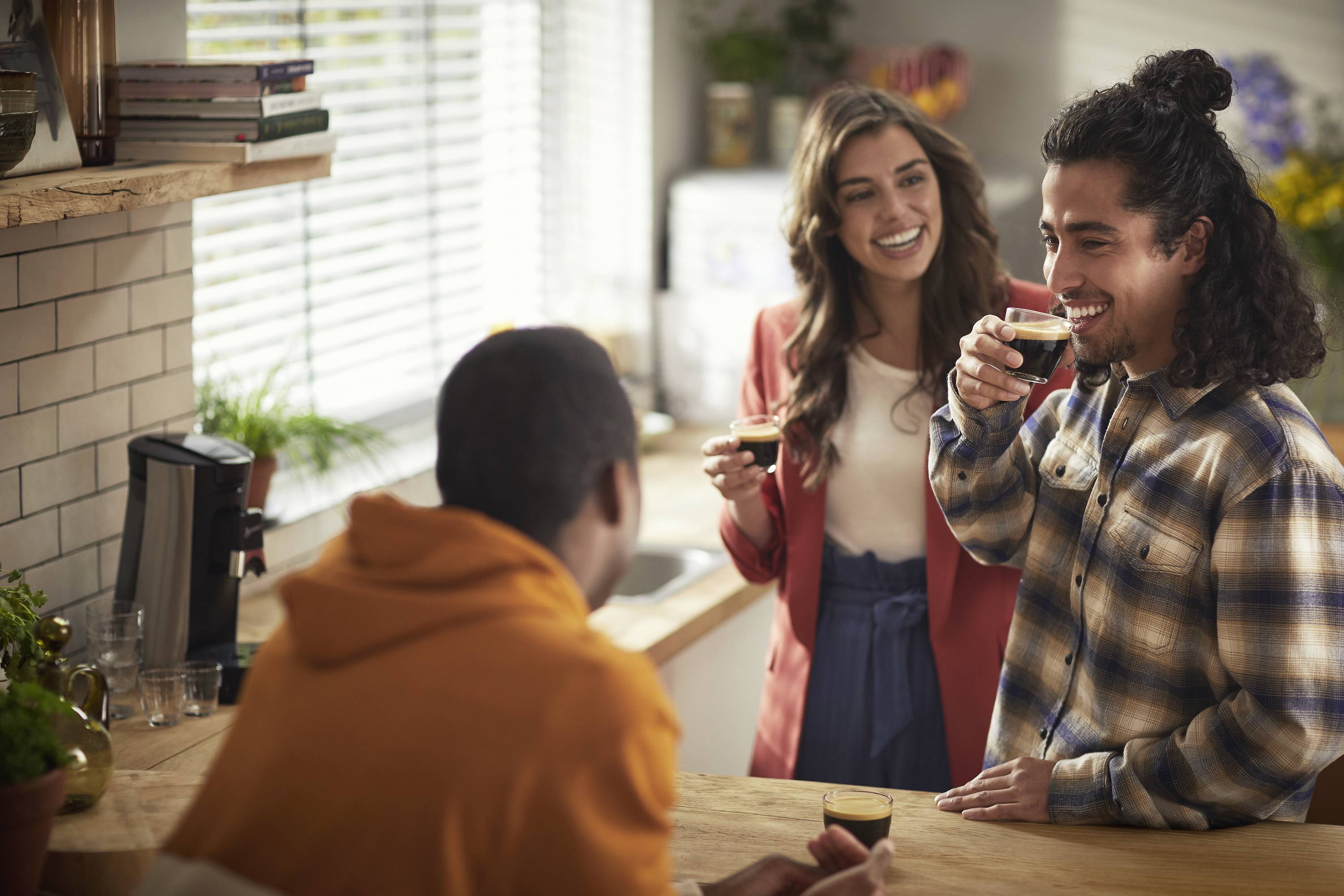Trois personnes dans une cuisine, riant et discutant ensemble autour de boissons. L'atmosphère est chaleureuse et détendue.