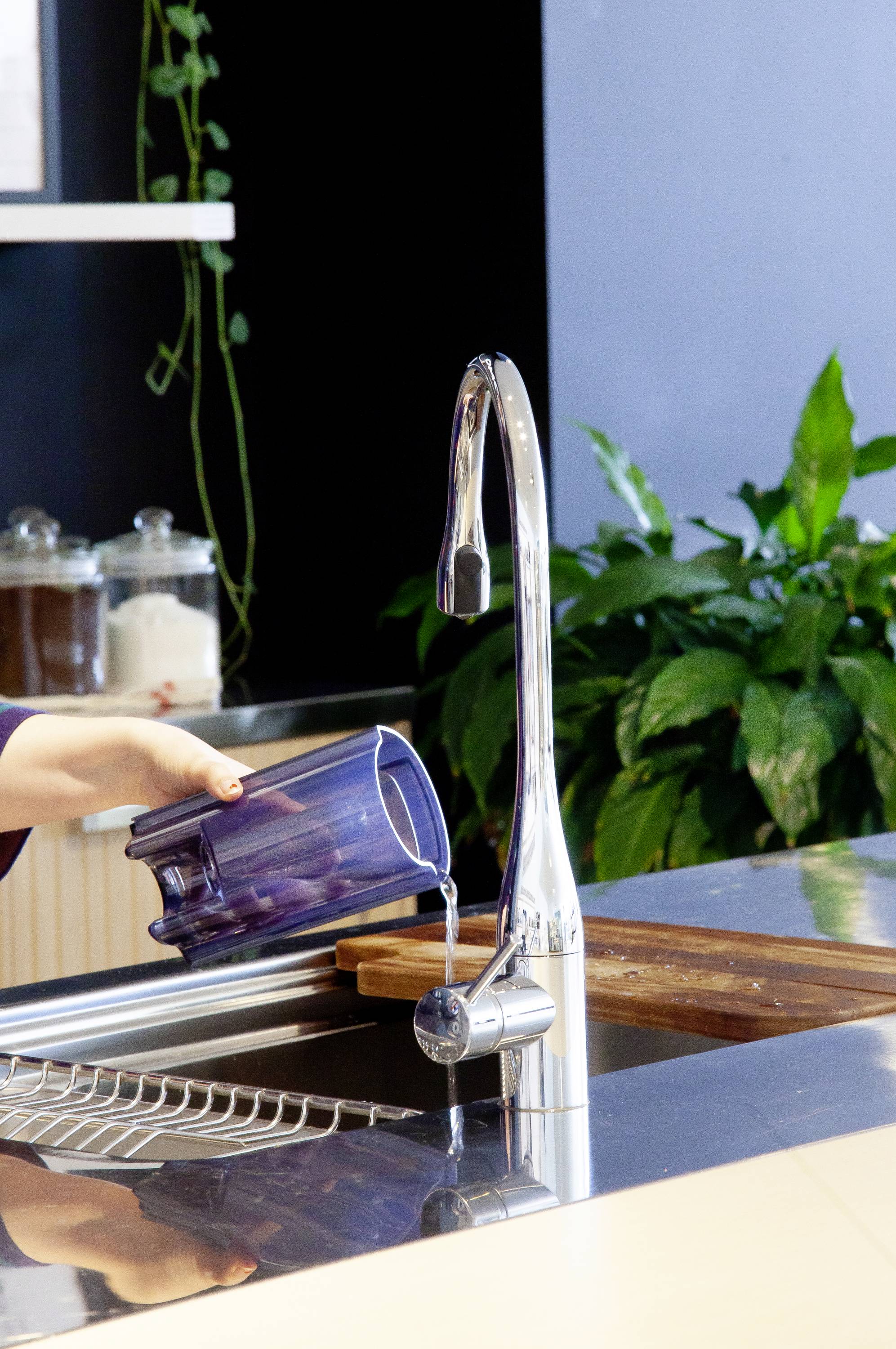 Une personne remplit un lavabo avec un broc bleu. Des décorations végétales et des ustensiles de cuisine sont visibles en arrière-plan.