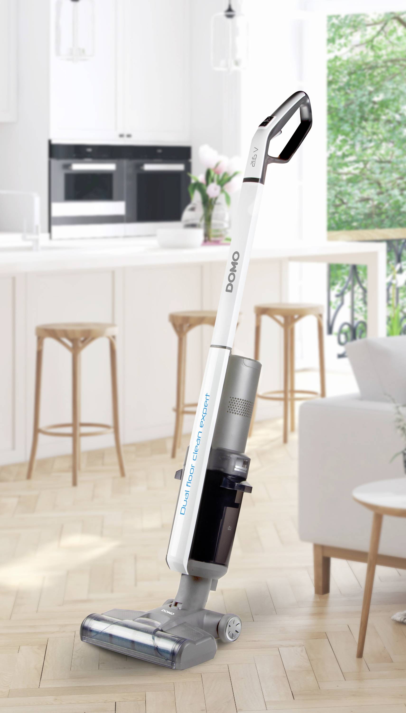 Un aspirateur blanc sans fil est posé sur un sol en bois clair dans une cuisine moderne aux meubles blancs et dotée de grandes fenêtres.