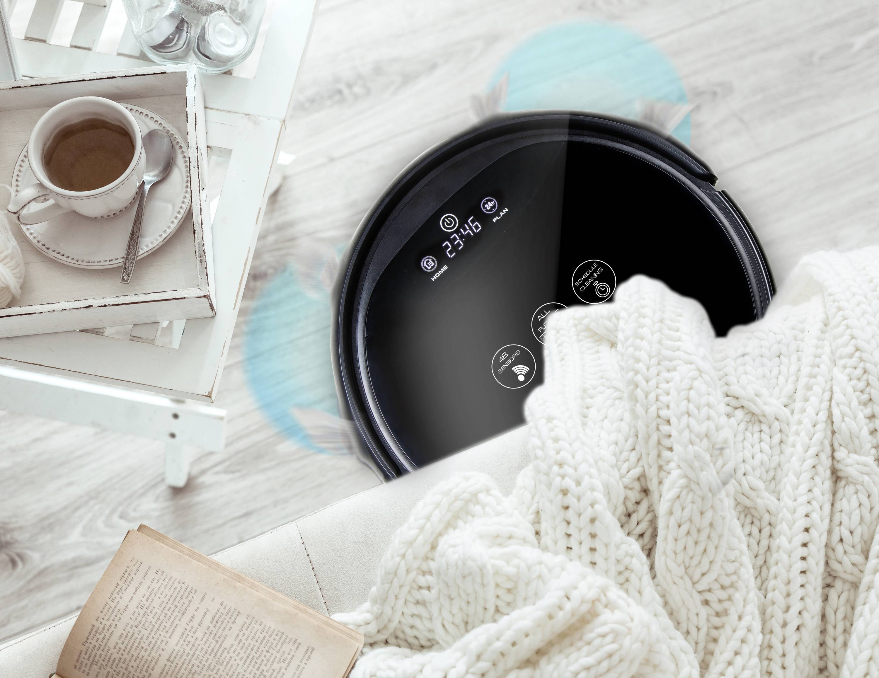 Un robot aspirateur noir nettoie le sol à côté d'une table avec une tasse de café et une couverture ouverte sur un fauteuil.