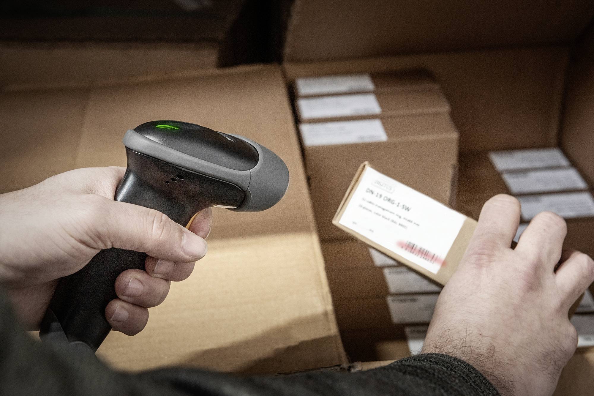 Une personne utilise un lecteur de codes à barres pour scanner une étiquette sur un carton. D'autres cartons sont visibles en arrière-plan.