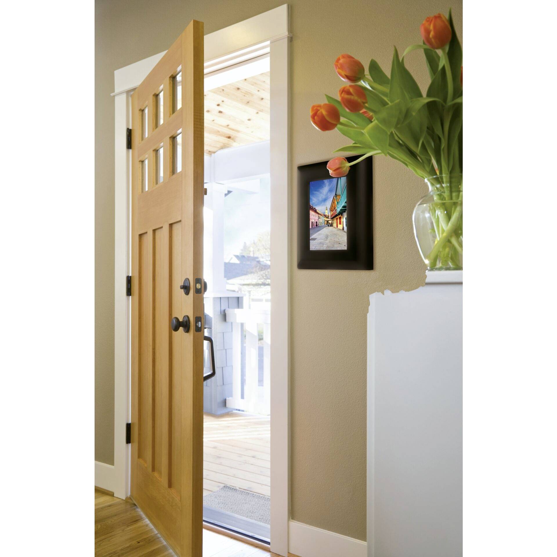 Une porte en bois ouverte avec un tableau accroché au-dessus, laissant apercevoir un balcon lumineux et ensoleillé. Un bouquet de fleurs sur la droite.