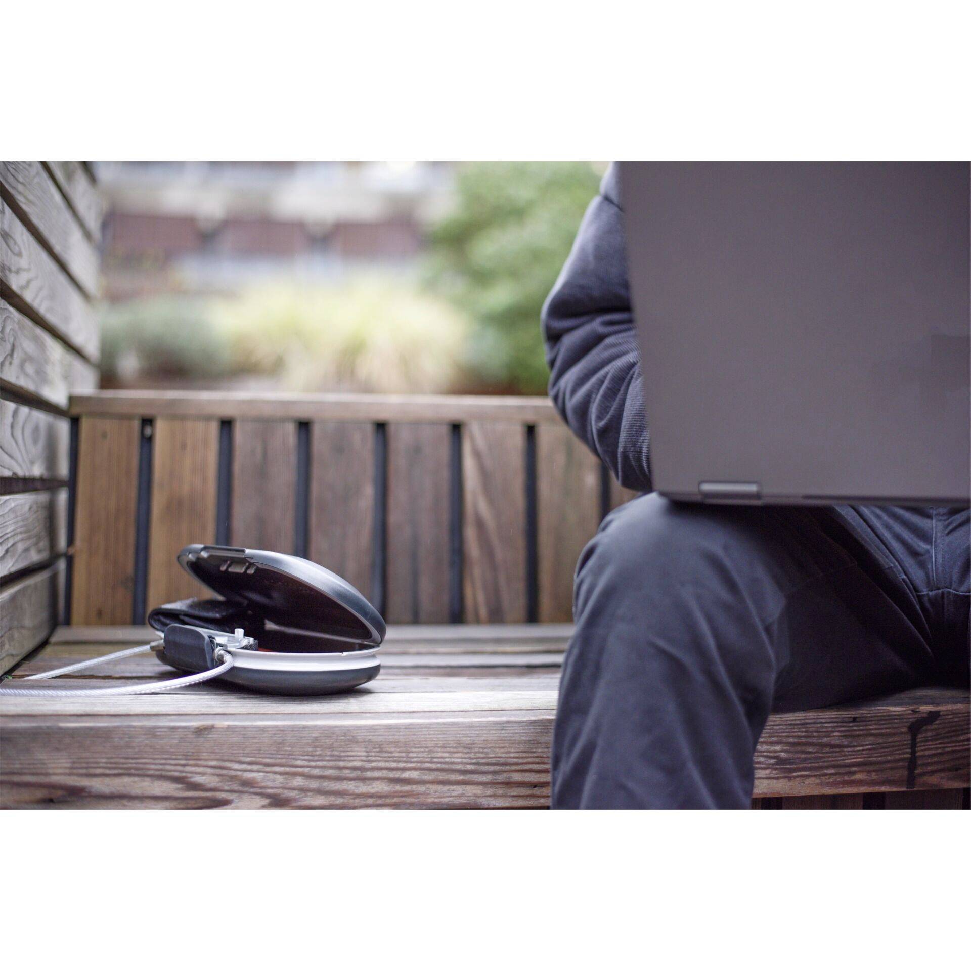 Une personne est assise sur un banc en bois avec un ordinateur portable sur les genoux. Des écouteurs sont posés à côté d'elle. L'environnement est flou.