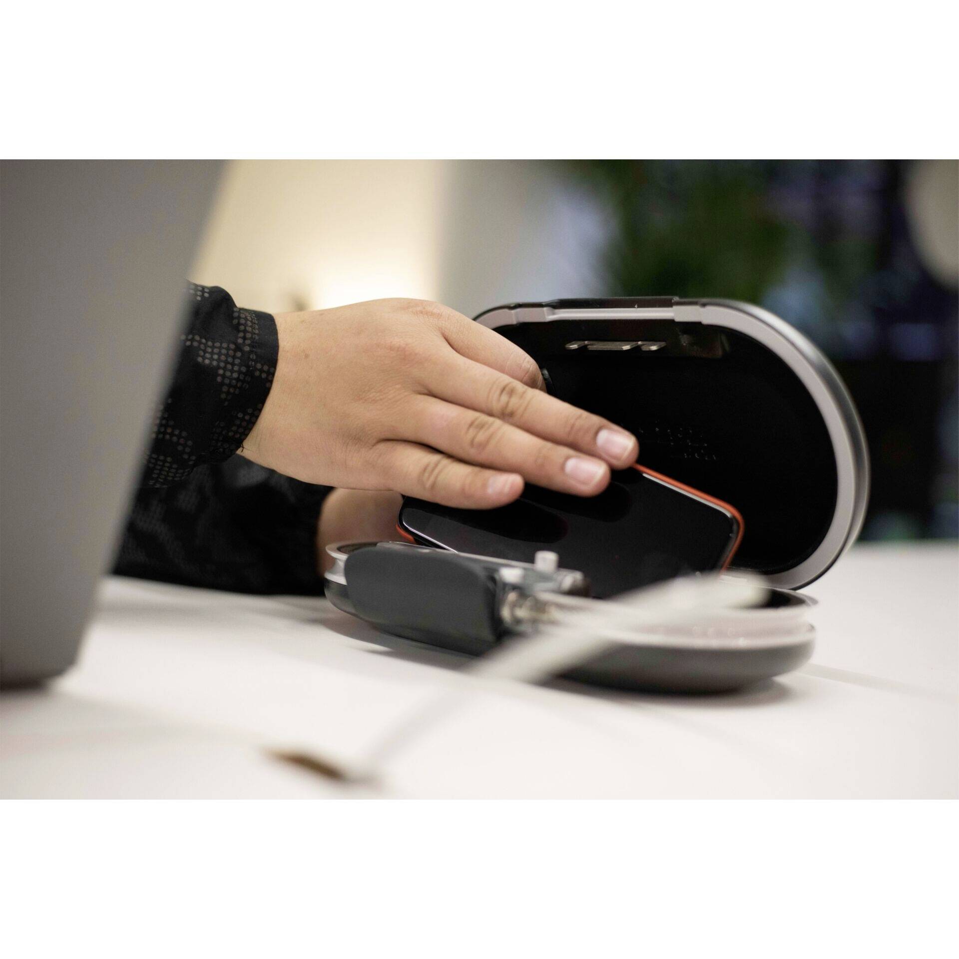 Une main pose un smartphone sur une station de charge sans fil posée sur une table. Un éclairage flou et un ordinateur portable sont visibles en arrière-plan.