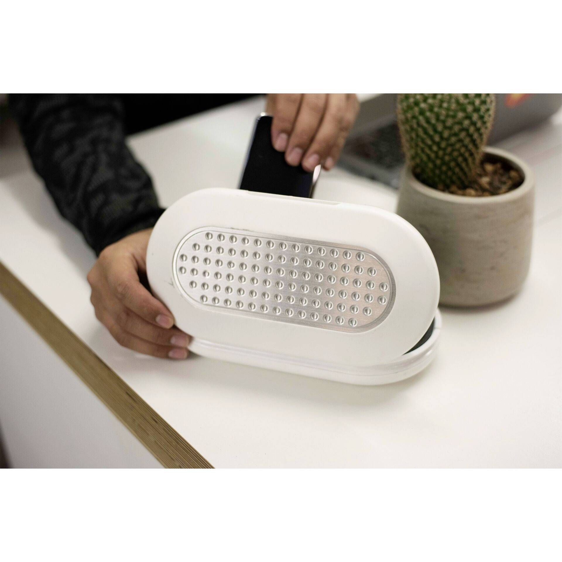 Une personne pose un smartphone sur un chargeur sans fil blanc, posé sur une table à côté d'un petit cactus dans un pot de fleurs.