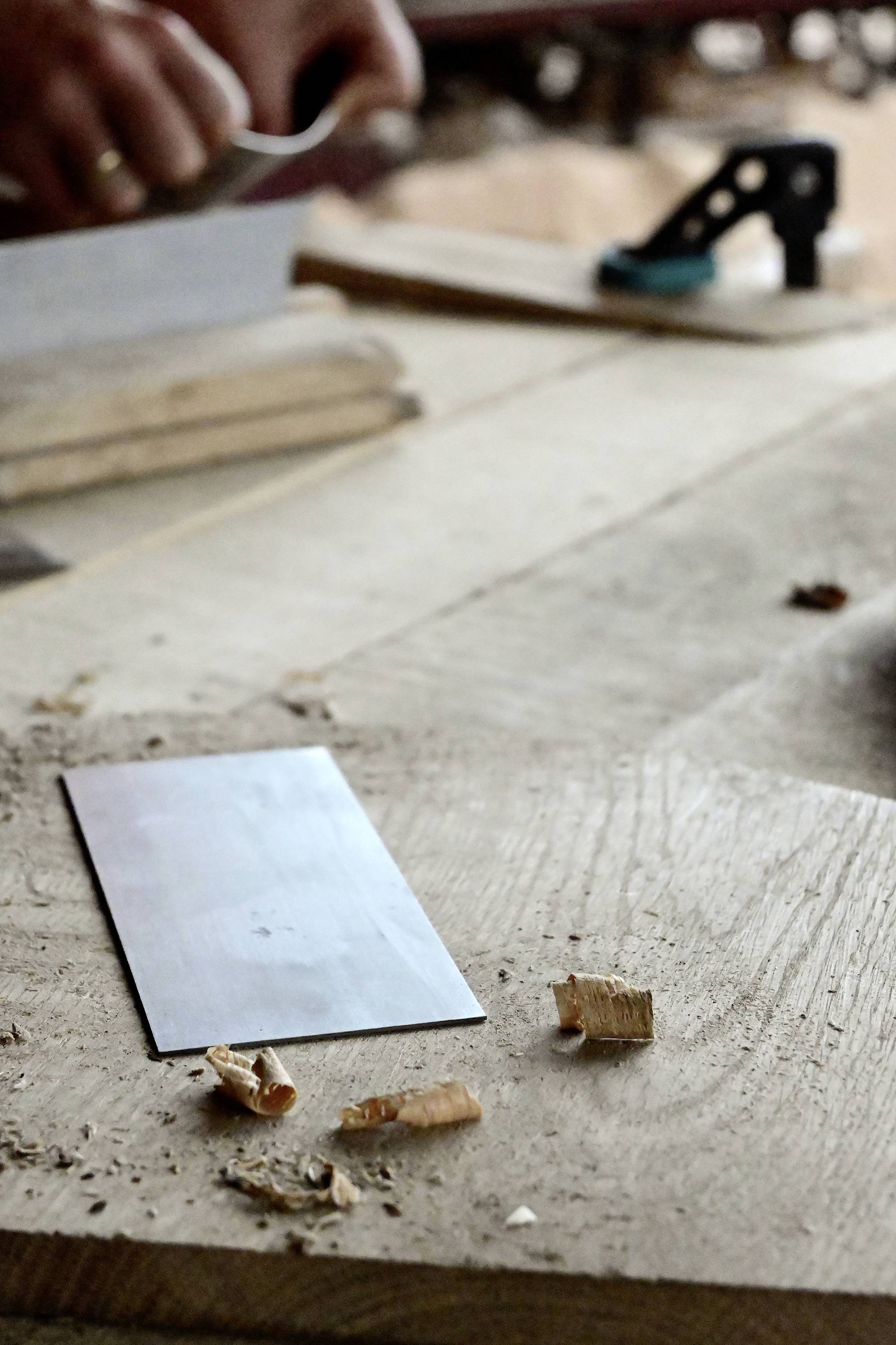 Un menuisier travaille le bois avec un rabot sur un établi. Des copeaux de bois sont éparpillés, un outil métallique est posé au premier plan.