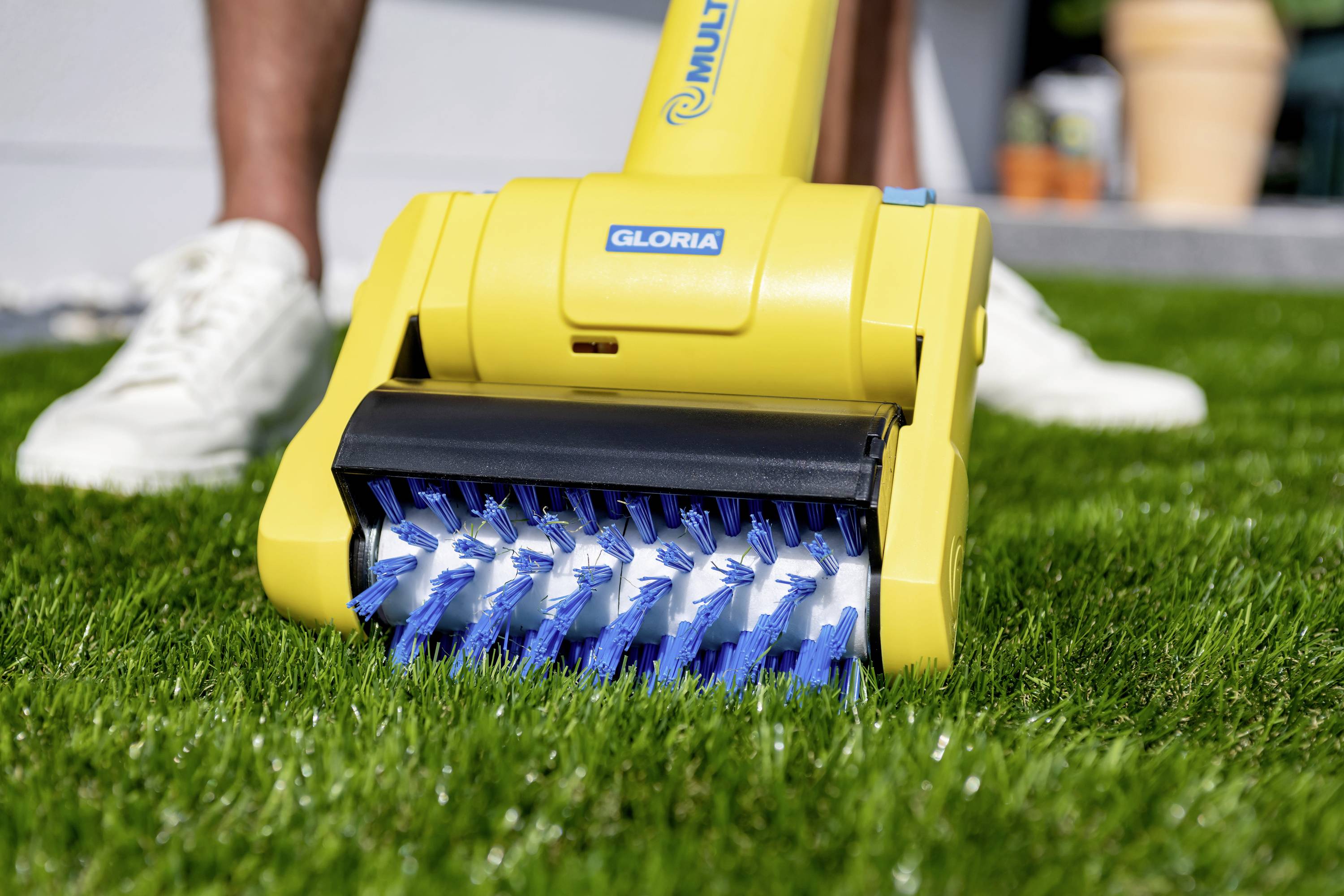 Une personne utilise un appareil de jardinage électrique jaune avec des brosses bleues pour entretenir la pelouse. Des jambes portant des chaussures blanches sont visibles en arrière-plan.