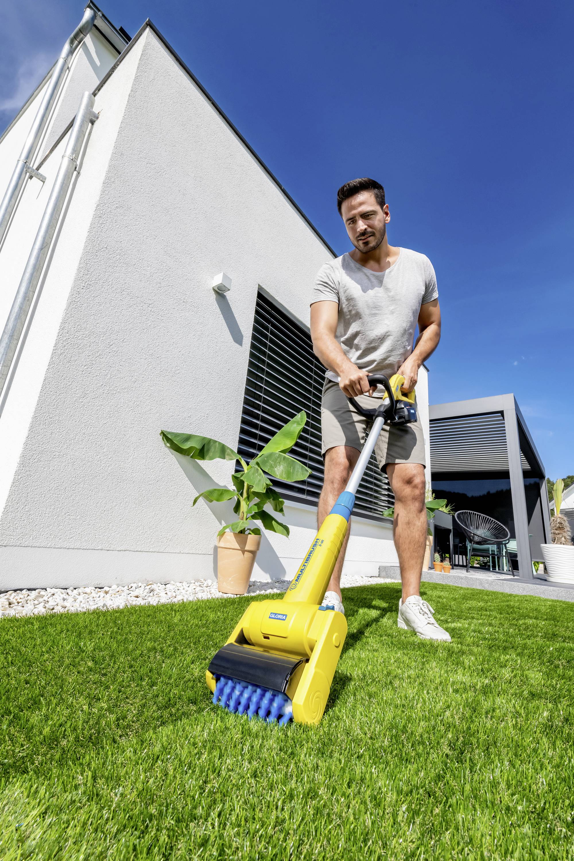 Un homme entretient la pelouse d'une maison moderne avec un appareil de nettoyage jaune. En arrière-plan, on aperçoit des plantes et un ciel bleu.